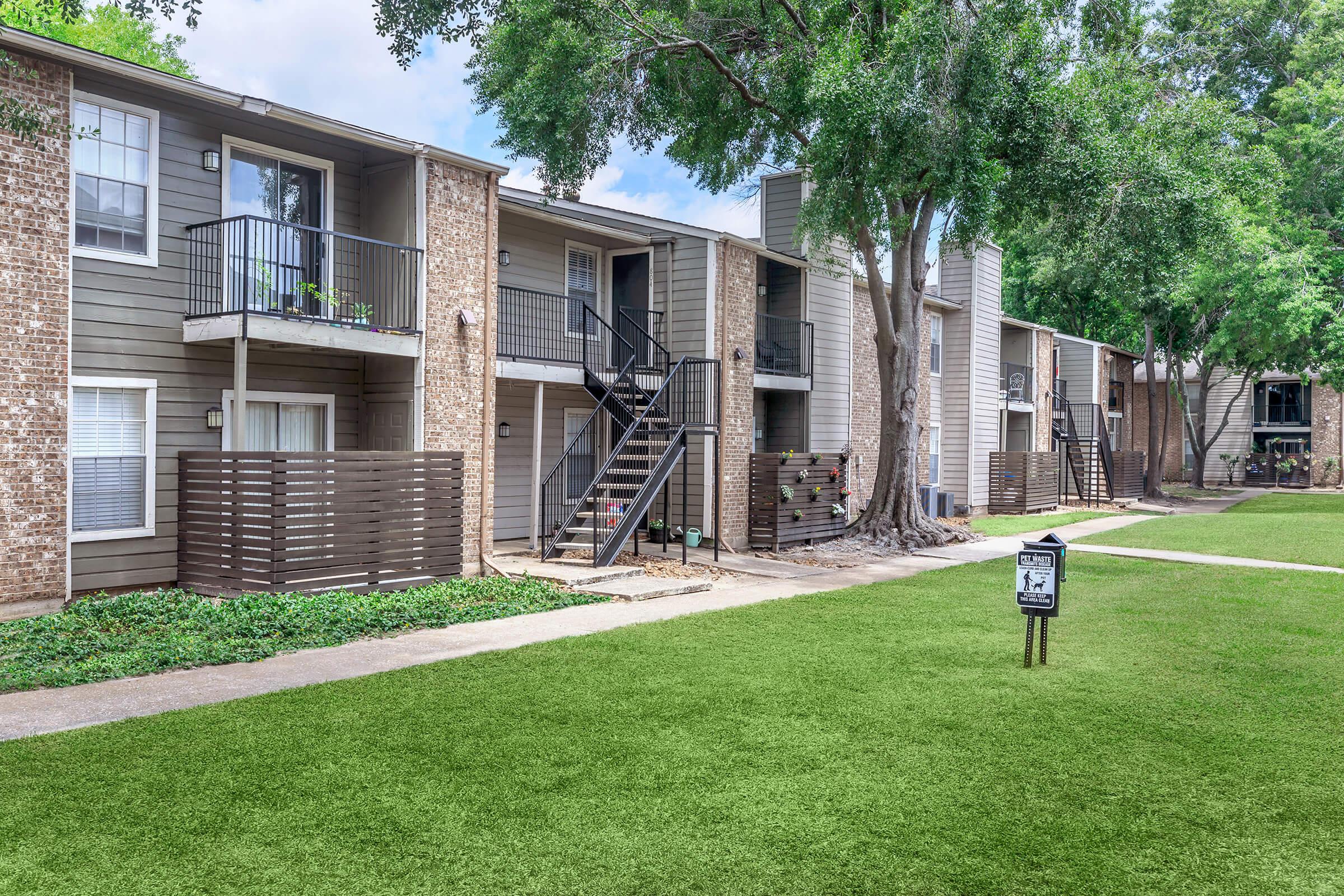 A view of an apartment complex featuring several two-story buildings with balconies. The buildings have a mix of brick and siding exteriors. A grassy area is in the foreground, and a sign is visible on the lawn. Trees provide shade along the pathway between the buildings.