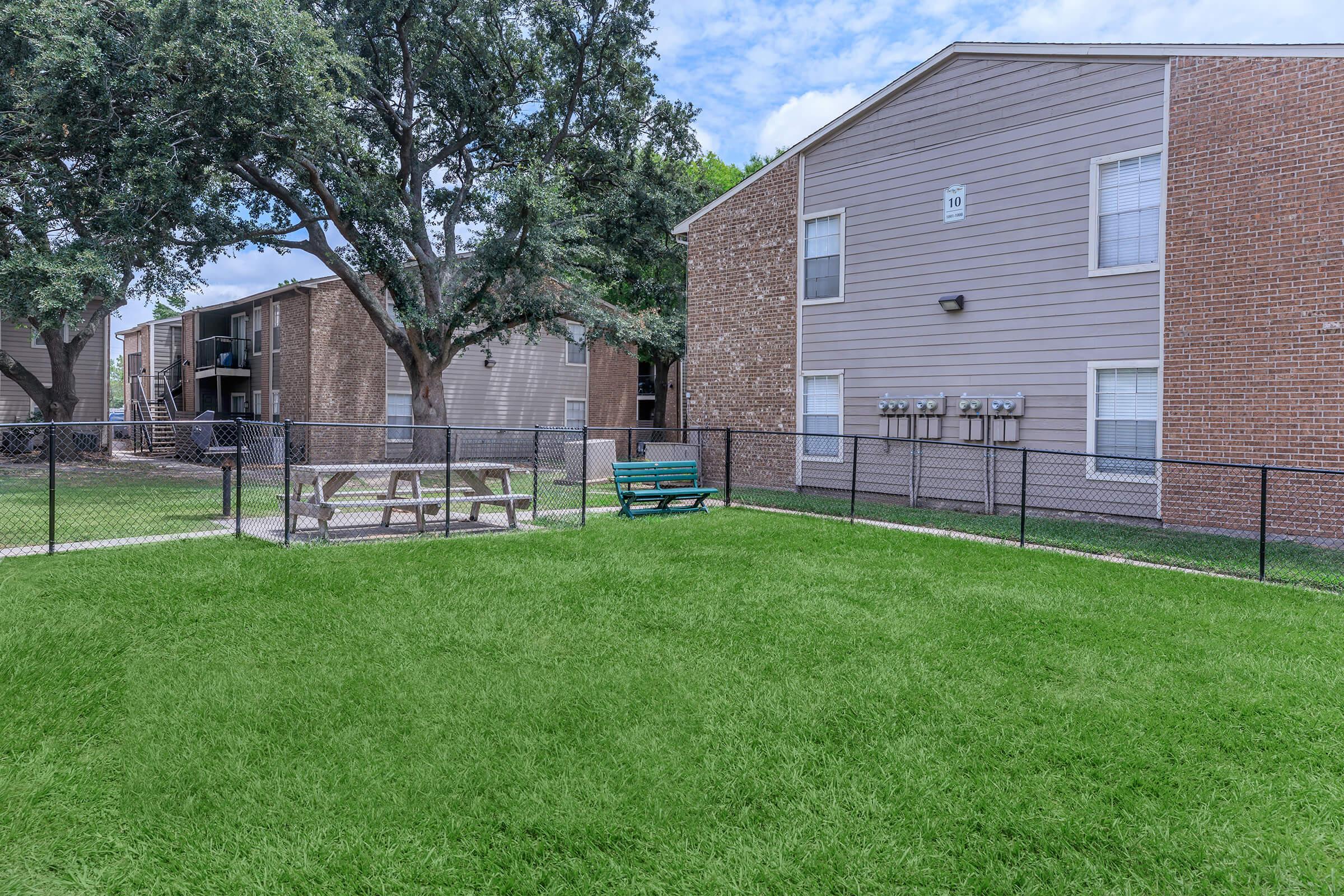 A view of an apartment complex with two buildings visible. In the foreground, there is a fenced-in grassy area featuring a picnic table and a green bench. Trees provide some shade, and the sky is partly cloudy. The scene conveys a quiet outdoor space for residents to relax.