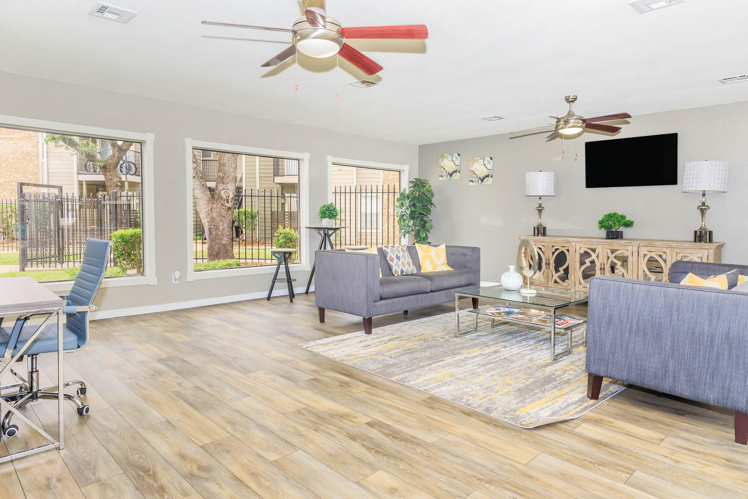 Bright and modern living room featuring two gray sofas with yellow and white accents, a coffee table, and a decorative side table. Large windows provide natural light and views of greenery outside. Ceiling fans add comfort, and a stylish console table with decorative items enhances the decor.