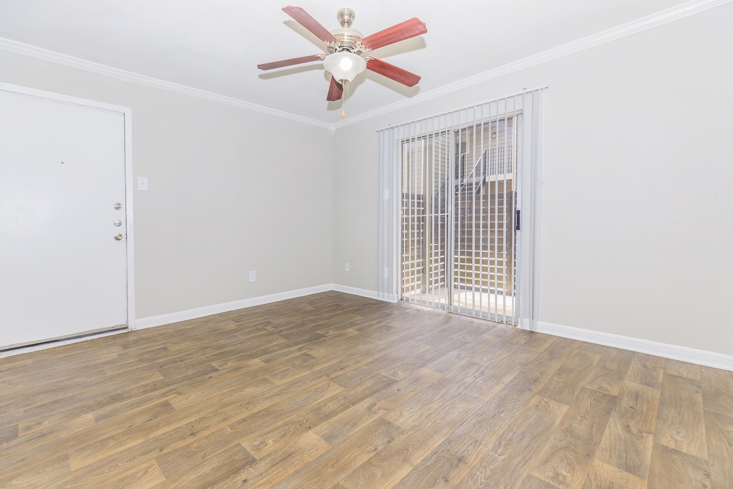 Bright, empty living room featuring light-colored walls and a ceiling fan with red blades. There are large sliding glass doors leading outside, and a simple front door on one side. The floor is covered with wood-like laminate flooring, enhancing the open and airy atmosphere.