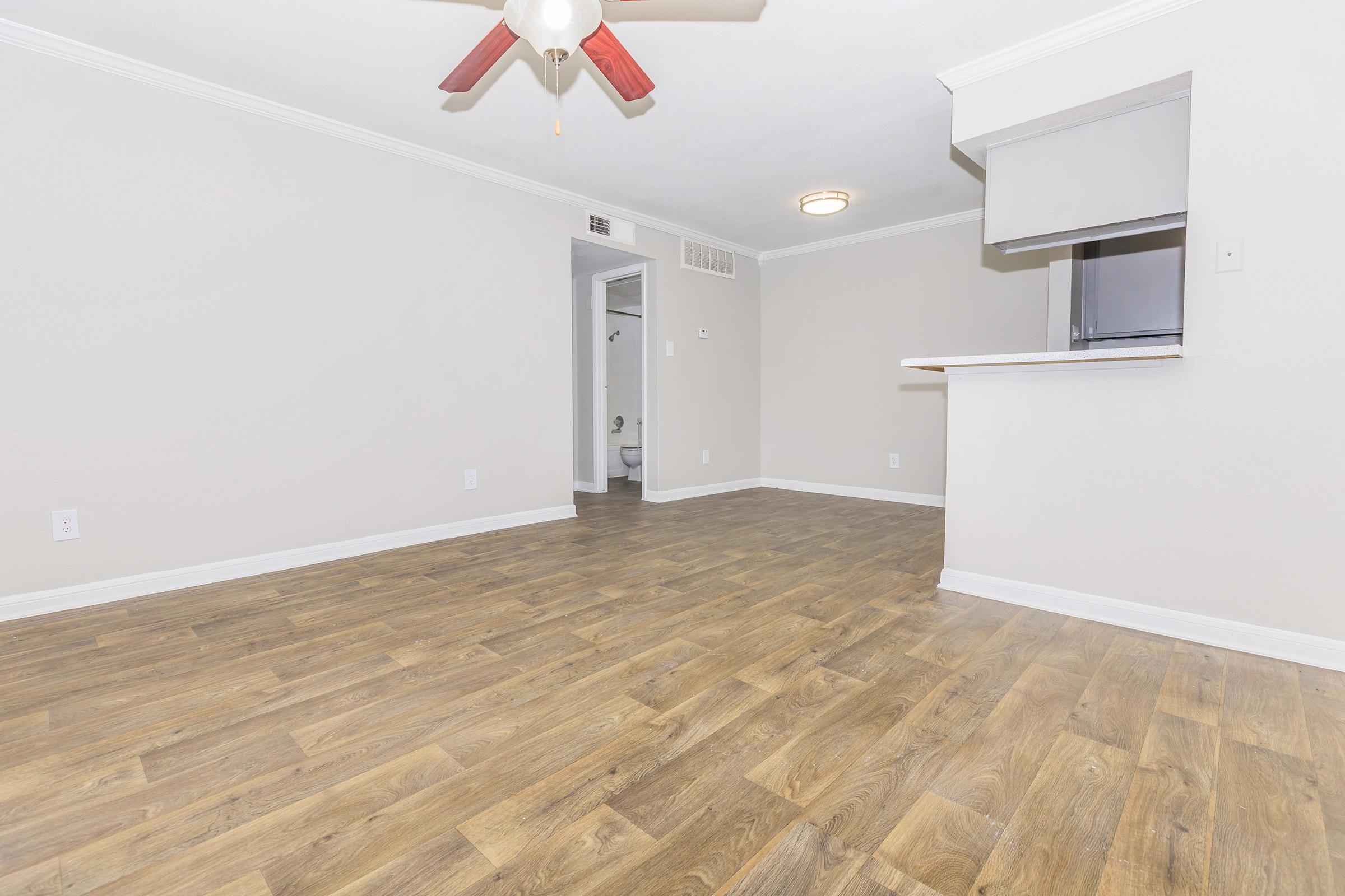 Empty room with light-colored walls and a wood laminate floor. A ceiling fan is mounted in the center. To the left, an area for a small kitchen counter is visible. A doorway leads to a bathroom in the back. The space is well-lit and appears freshly painted, creating a clean and inviting atmosphere.