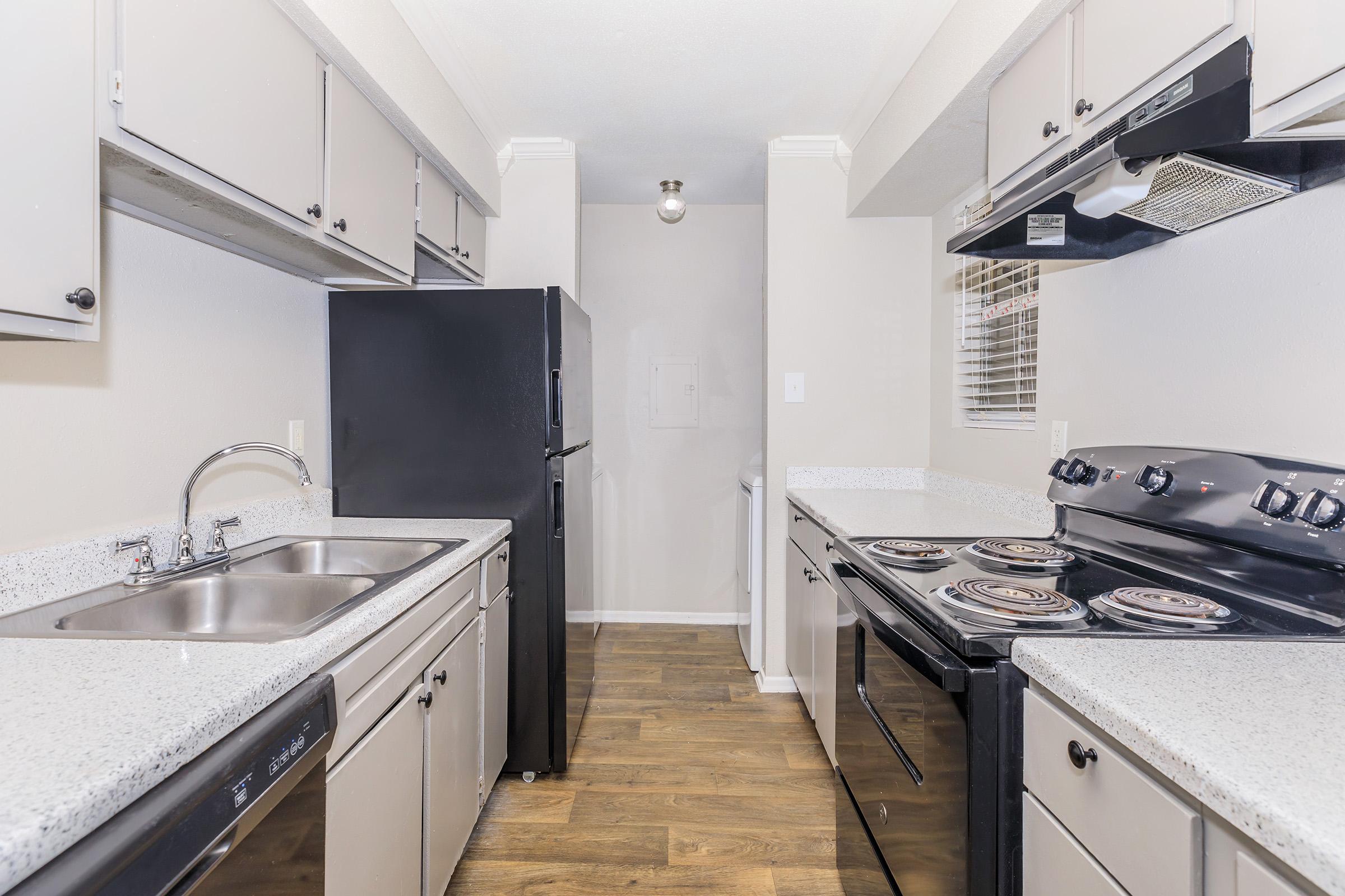 A modern kitchen featuring a black refrigerator, double sink, and stove with an overhead microwave. The cabinetry is light grey, and the countertops are a speckled white. The flooring is a warm wood tone, and the space is well-lit, creating a clean and inviting atmosphere.