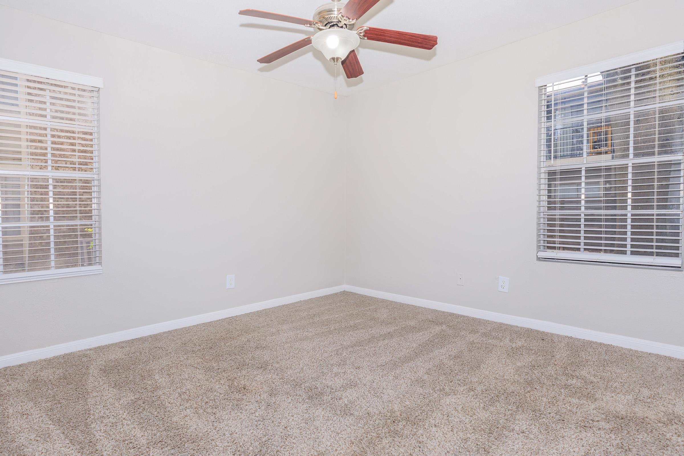 A vacant room with light-colored walls and beige carpet. There are two windows with white blinds on opposite walls, allowing natural light to enter. A ceiling fan with wooden blades hangs from the ceiling, creating a bright and airy atmosphere.