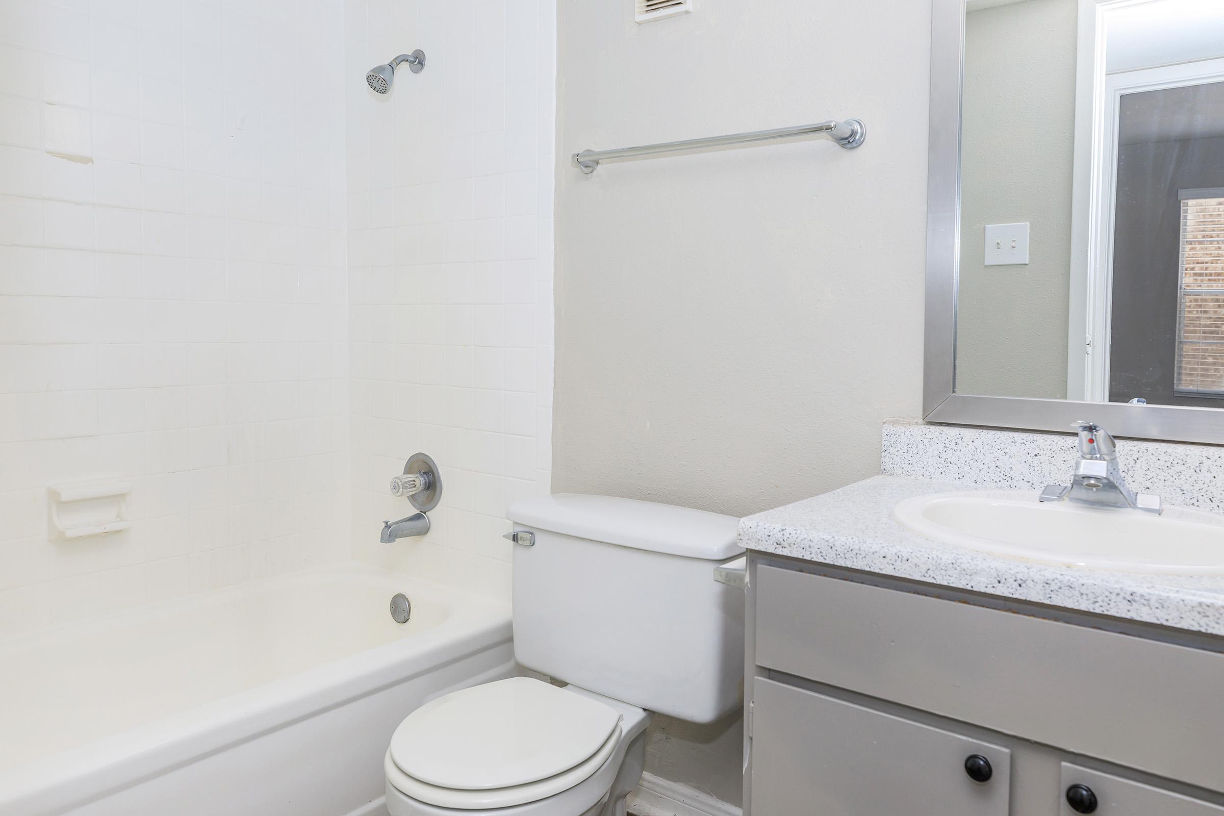 A clean and simple bathroom featuring a white bathtub, a toilet, and a sink with a countertop. The walls are light-colored, and there is a mirror above the sink. A towel bar is mounted on the wall, and the bathroom appears well-lit with natural light coming from a nearby window.