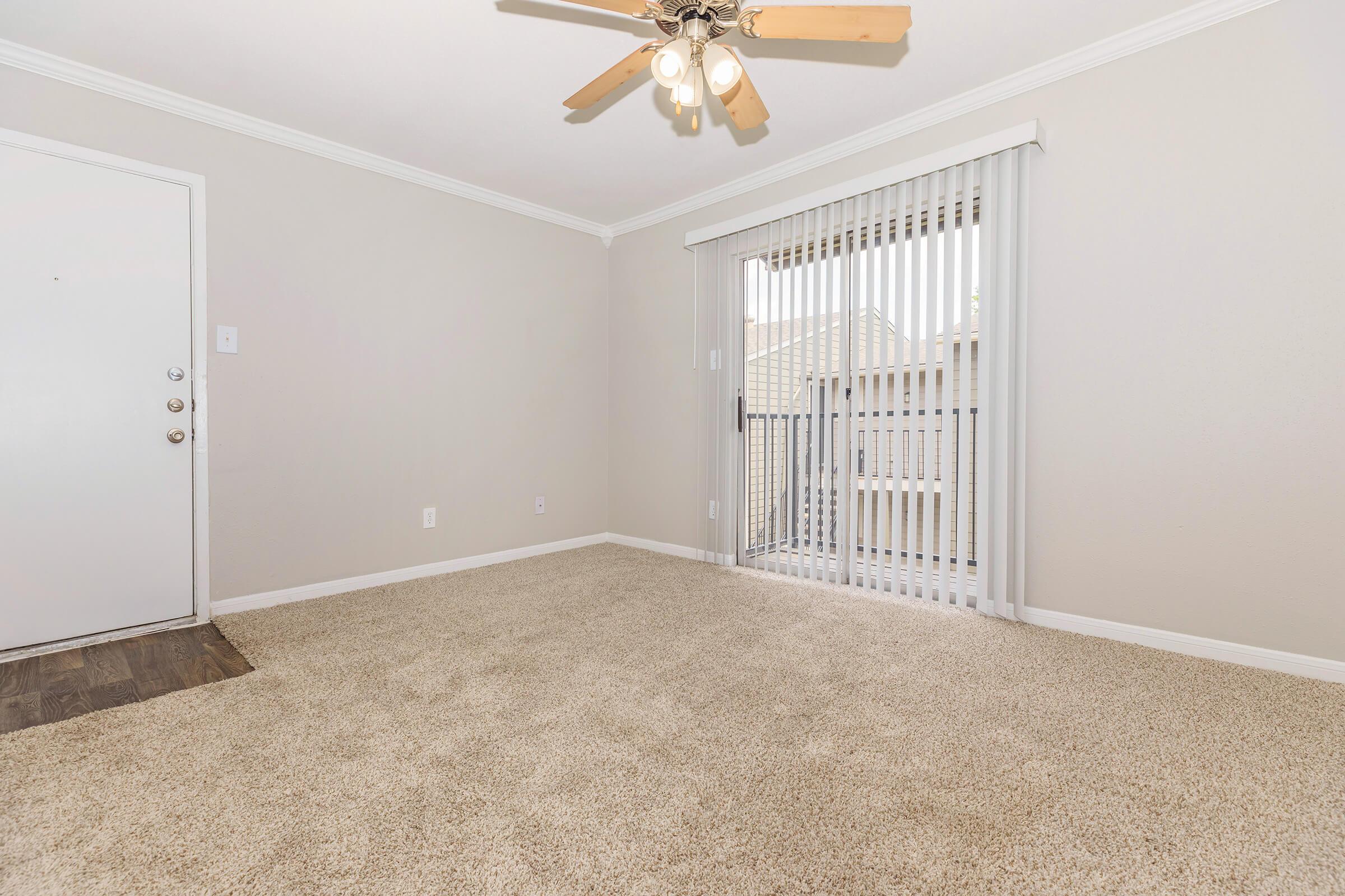 A well-lit, empty living room with beige carpet and light-colored walls. A ceiling fan with wooden blades hangs from the ceiling. There's a sliding glass door with vertical blinds leading to a balcony, and a front door visible on the left side of the image. The space feels open and inviting.