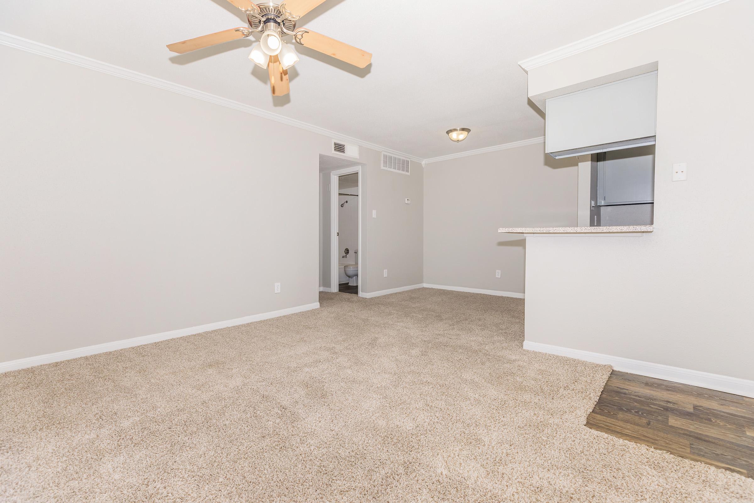 A spacious, carpeted living area featuring a ceiling fan, light-colored walls, and a bar counter leading to a kitchen area. There is a glimpse of a bathroom in the background through an open doorway. The flooring transitions from carpet to a wood-like surface near the bar.