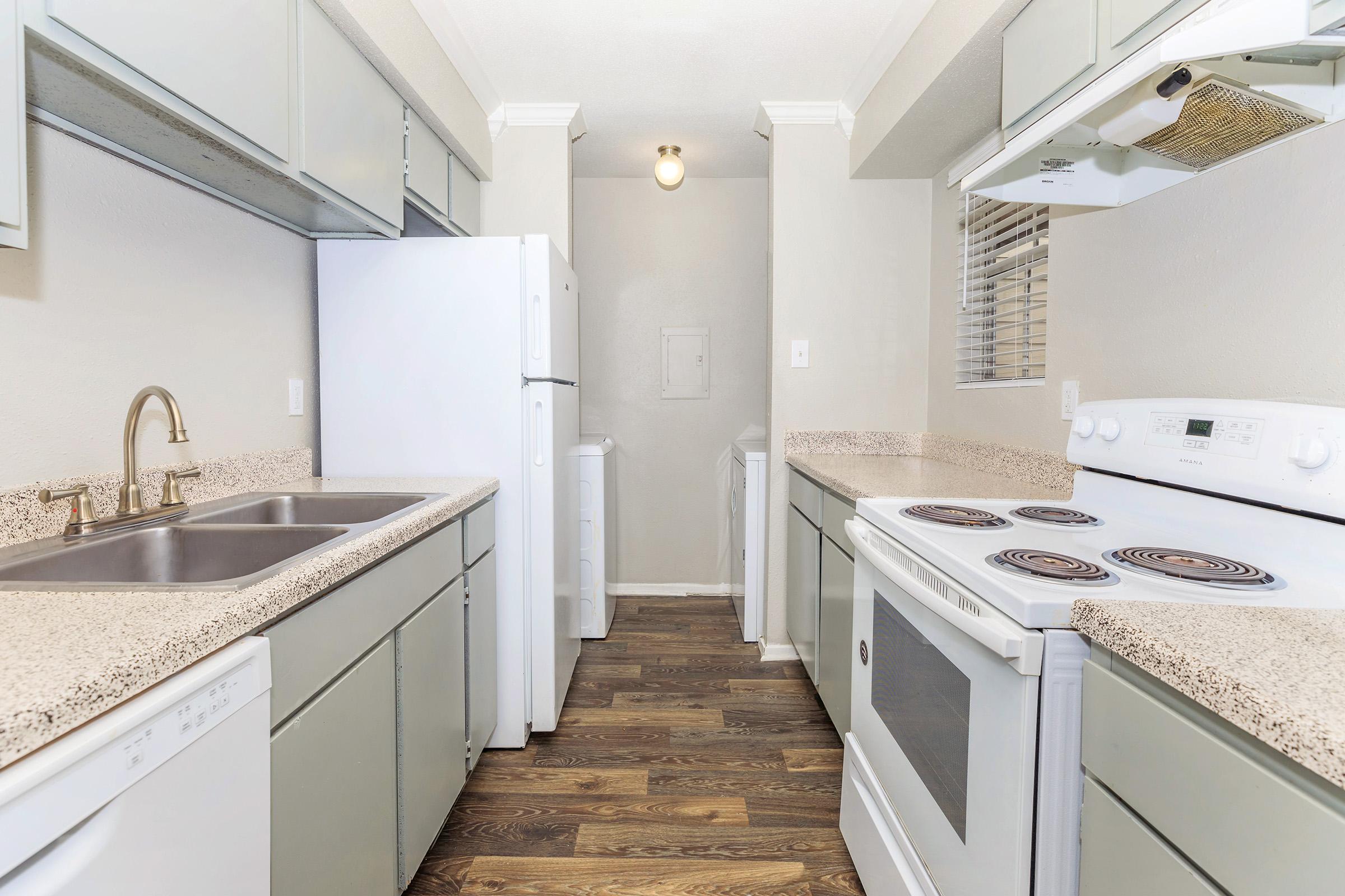 A modern kitchen featuring light gray cabinets, a double sink, a white refrigerator, and an electric stove. The countertop is a speckled beige design, and the flooring is dark wood. The space is well-lit with overhead lighting and has a clean, minimalist look.