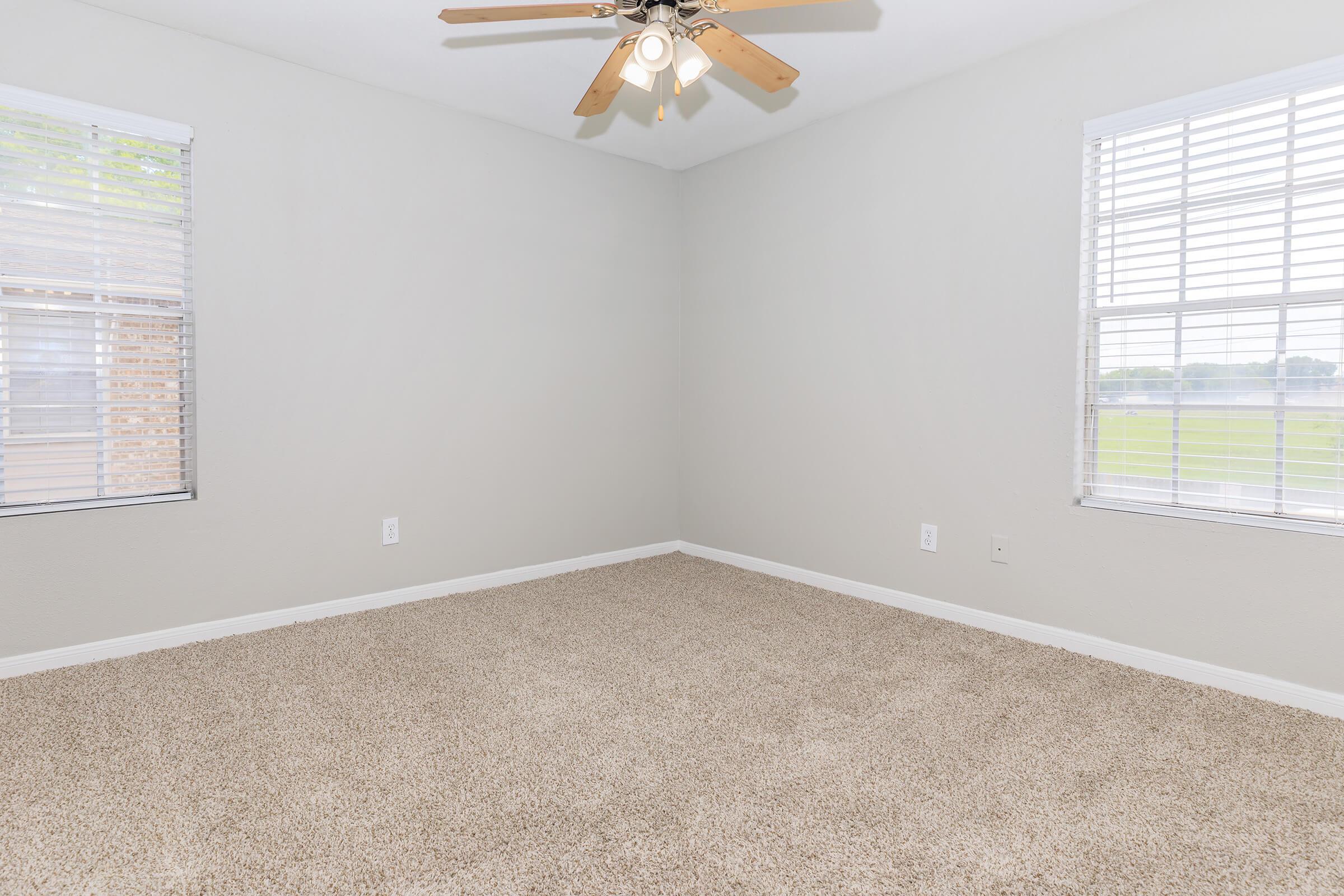 A clean, empty room featuring beige carpet, light gray walls, and two windows with white blinds. A ceiling fan with wooden blades hangs from the ceiling. The space is well-lit and has a bright, airy feel.