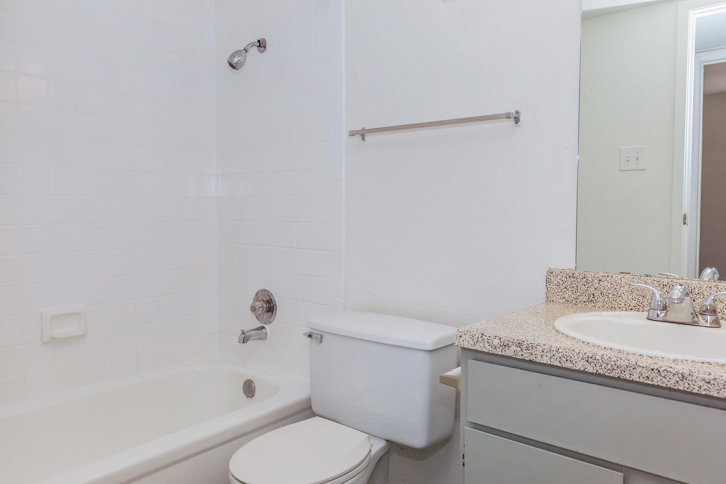 A clean and simple bathroom featuring a white bathtub with a chrome showerhead, a toilet, and a sink with a granite countertop. The walls are tiled in white and the space is well-lit, creating an airy atmosphere. A towel rack is mounted on the wall beside the sink.