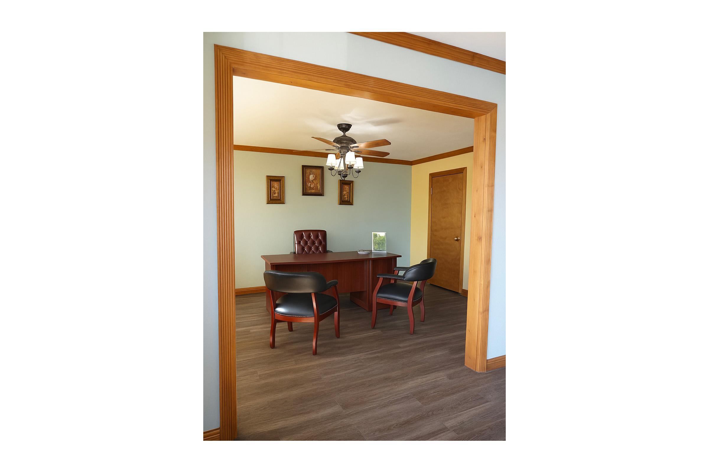 A well-lit office space featuring a wooden desk with a red upholstered chair, two black chairs in front, and wooden flooring. The walls are painted blue with wooden trim, and there are two framed pictures hung above the desk. A ceiling fan is visible, adding to the light and airy atmosphere.