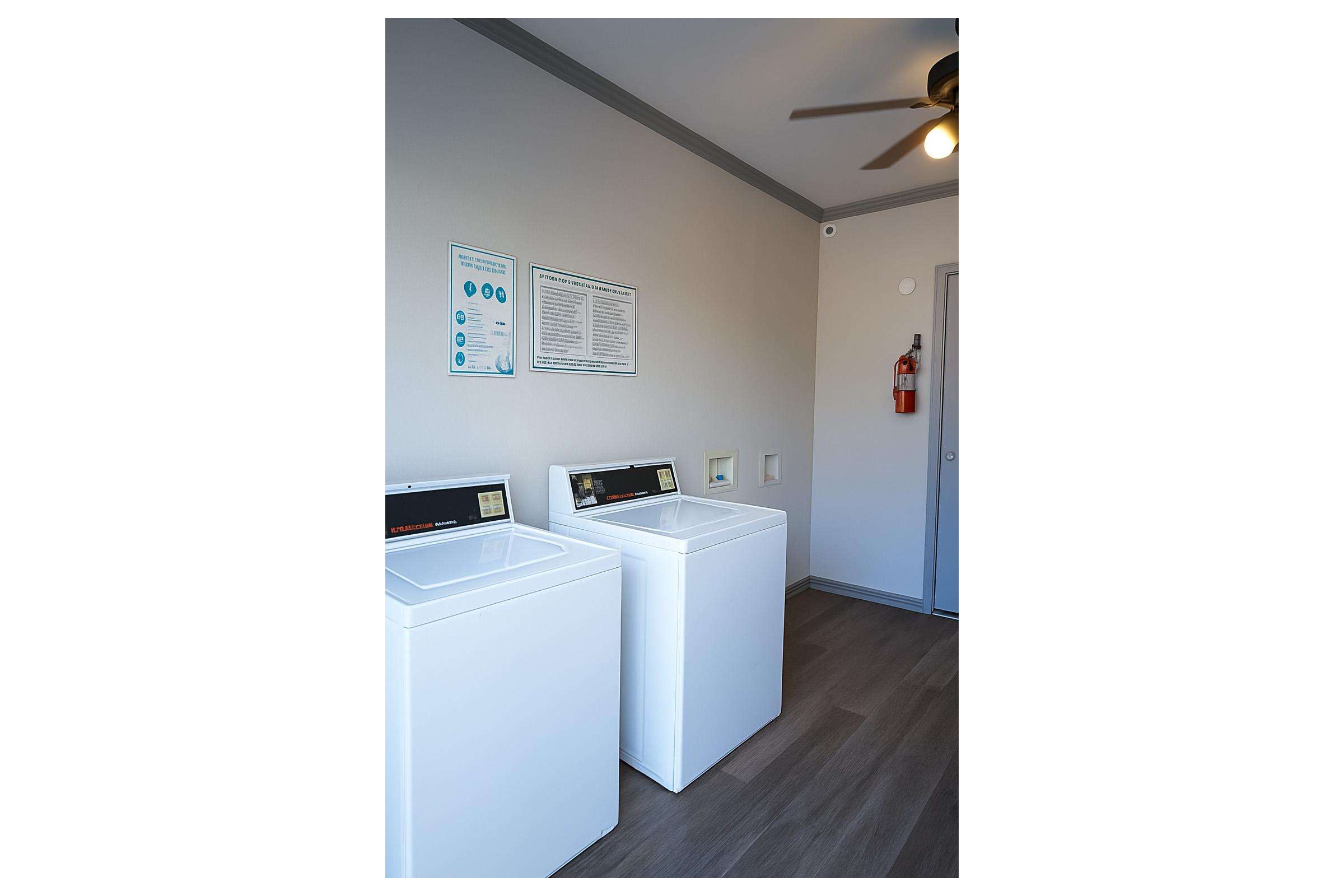 A clean laundry room featuring two white washing machines, a ceiling fan, a structured wall with a fire extinguisher, and informational posters. Natural light illuminates the space, highlighting the simple and functional design of the area.
