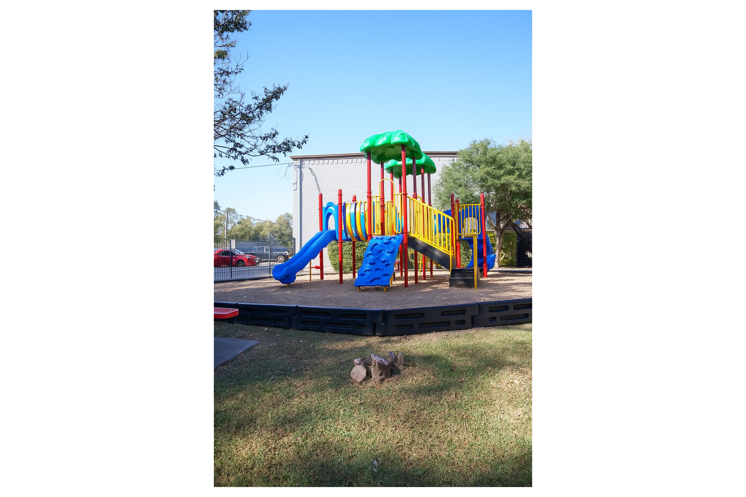 Brightly colored playground featuring slides, climbing structures, and tunnels, surrounded by grass and trees. The equipment is designed for children's play, with safety measures in place, against a clear blue sky. A paved area and some parked cars are visible in the background.
