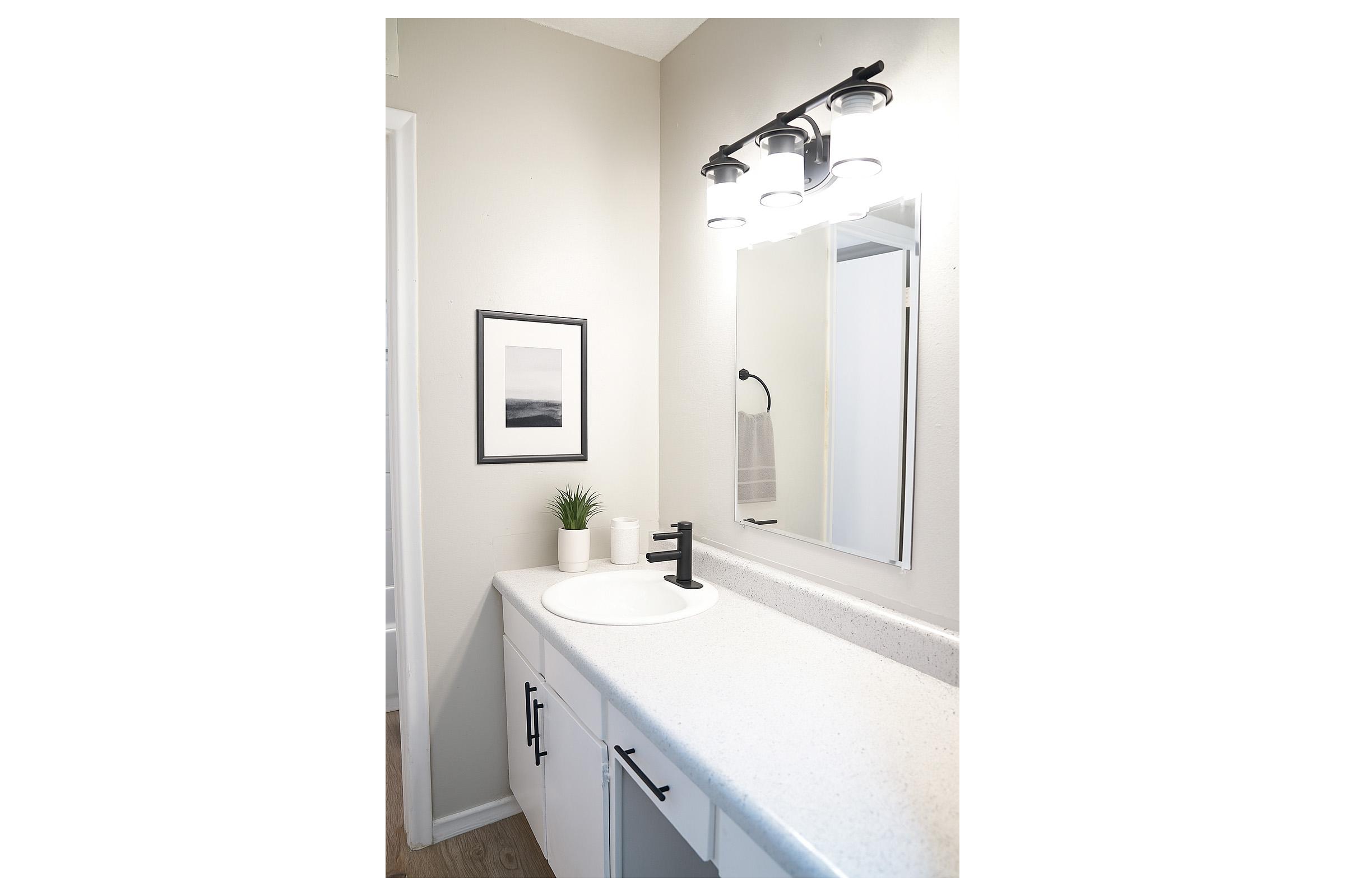 A modern bathroom featuring a white countertop with a round sink, a black faucet, and minimalist decor. A framed black-and-white photo is mounted on the wall, alongside a small potted plant and a white vase. The space is illuminated by stylish lighting above the mirror.