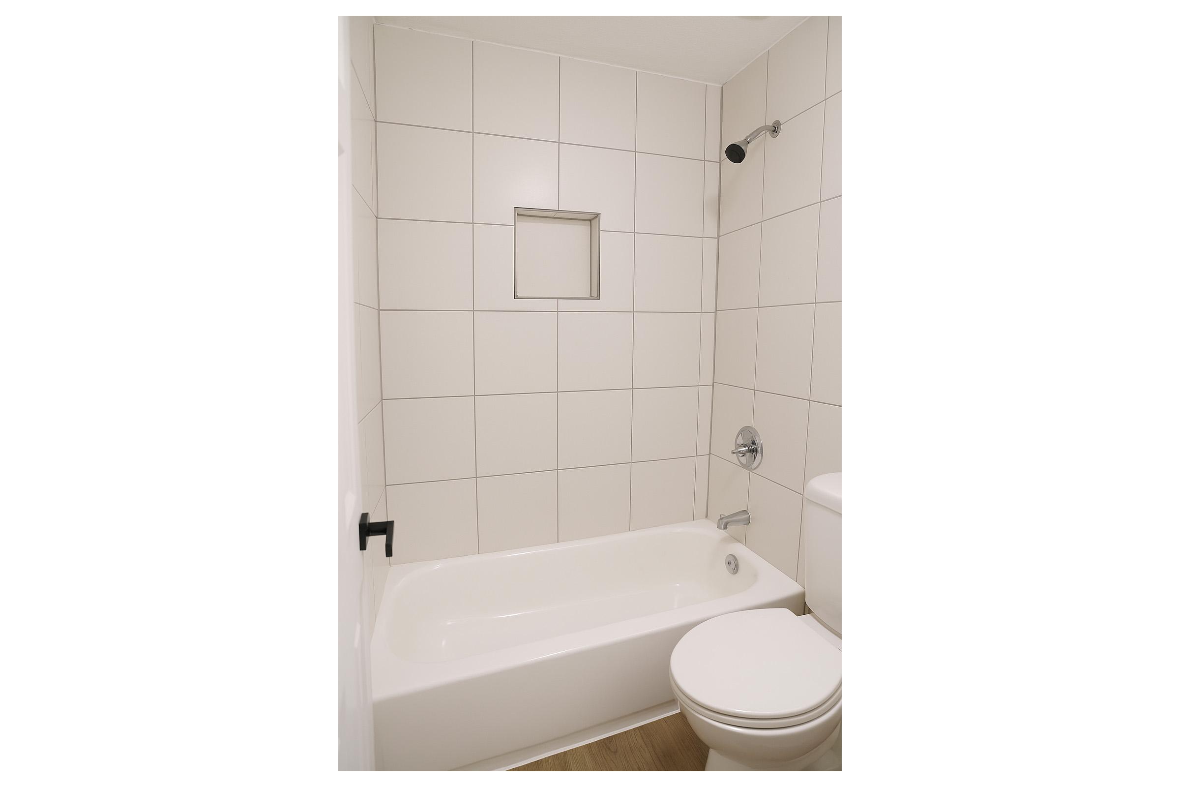 A modern bathroom featuring a clean, minimalist design. It includes a white bathtub with a showerhead, a small alcove for toiletries, and a white toilet. The walls are tiled with light-colored square tiles, and the floor is finished with wood-like flooring, creating a contemporary look.