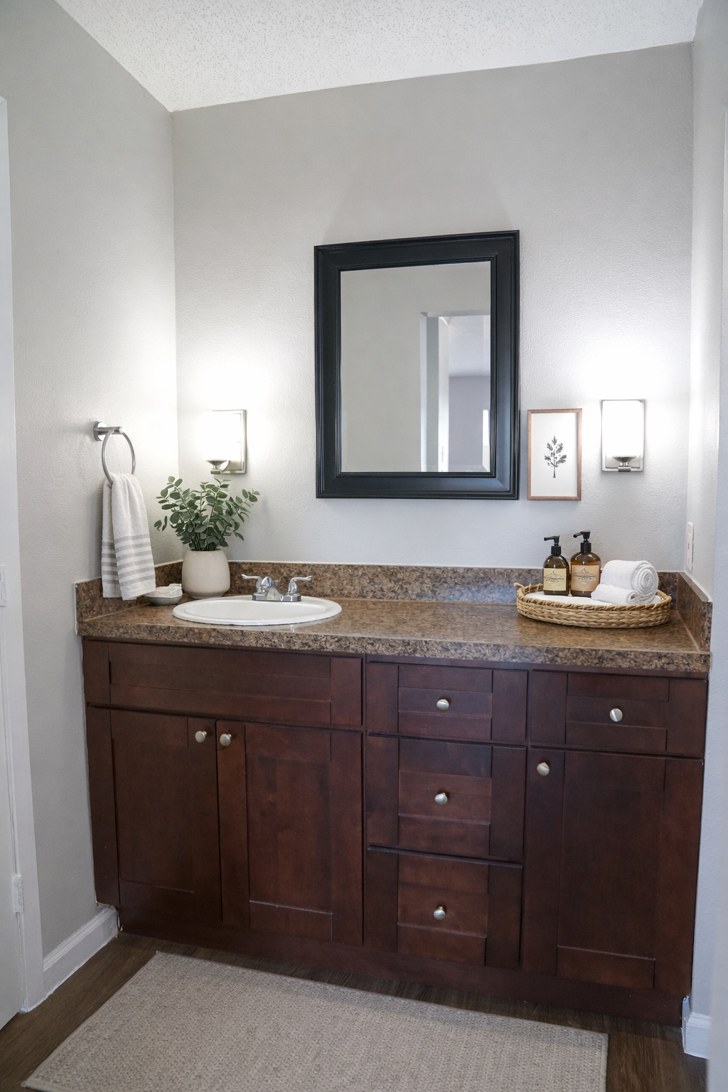 Modern bathroom with dark wooden cabinetry, a granite countertop, a round mirror framed in black, and elegant lighting. A small plant sits next to stylish soap dispensers on the countertop. A towel hangs on a wall hook, and a small basket with toiletries is placed nearby, enhancing the overall aesthetic.