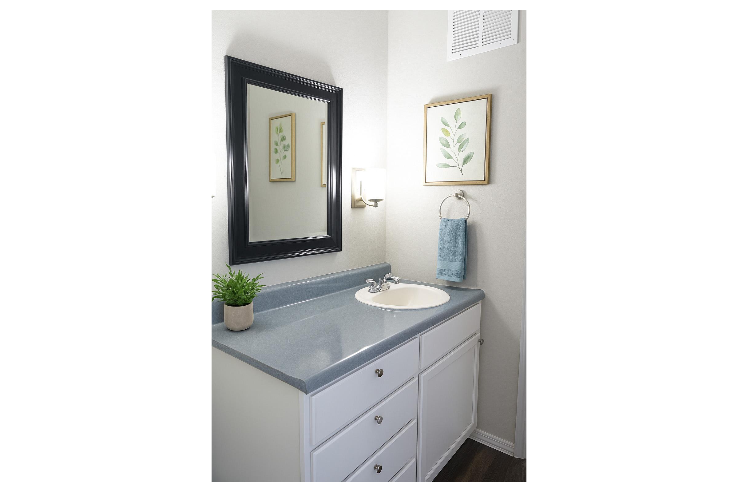 A clean and modern bathroom interior featuring a white vanity with gray countertop, a round sink, a wall-mounted light fixture, and a decorative mirror. There is a framed leaf print on the wall, a towel hanging from a ring, and a small potted plant on the countertop, creating a fresh and inviting atmosphere.