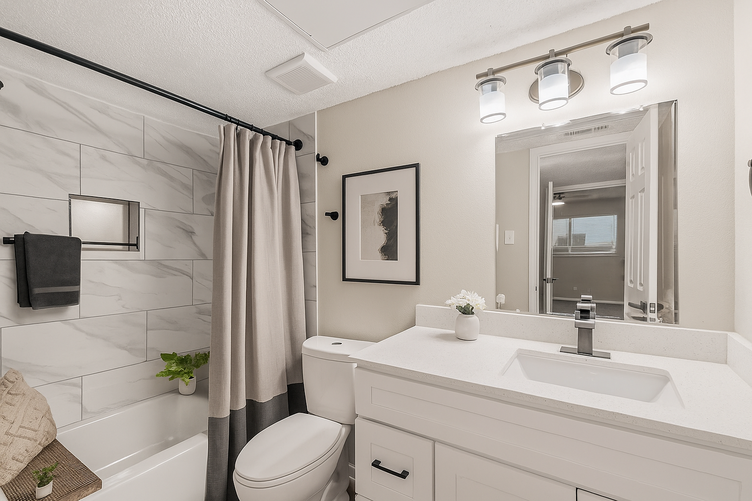A modern bathroom featuring a white vanity with a sink, a toilet, and a bathtub with a shower curtain. The walls are adorned with marble tiles, and there’s a contemporary light fixture above the mirror. Decorative elements include a plant and framed artwork, creating a clean and inviting space.