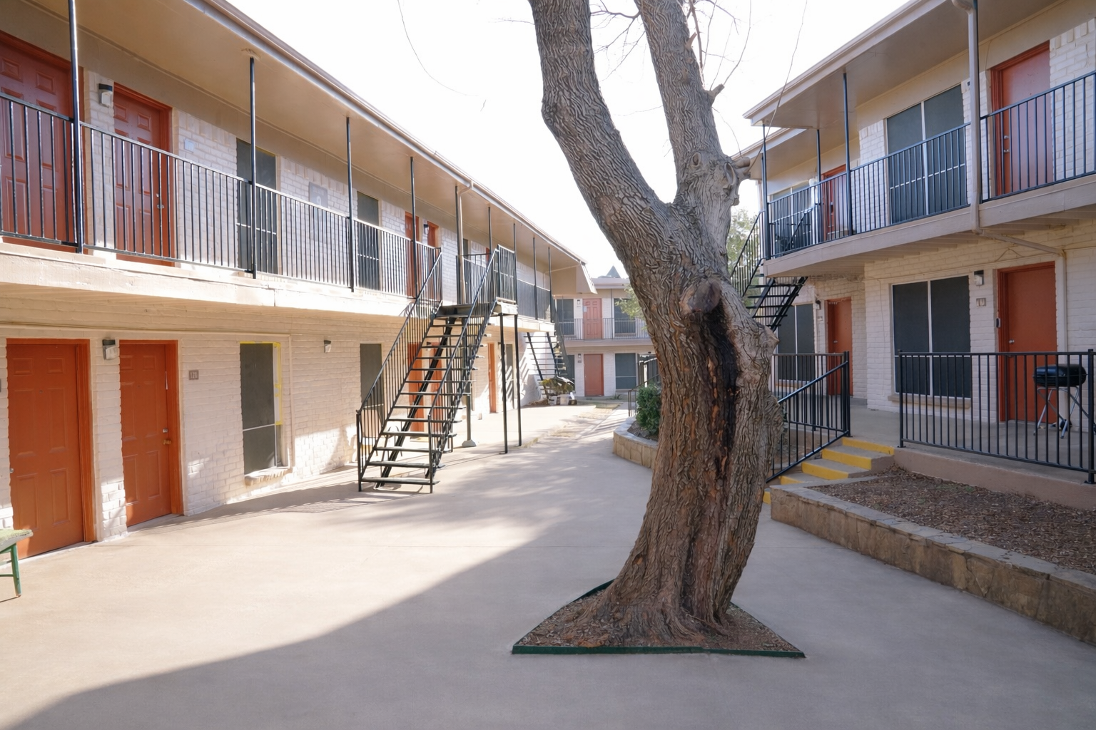 A view of a courtyard in a two-story apartment complex featuring several doors, staircases, and a central tree. The buildings have a light-colored exterior with black railings, and a paved walkway runs through the area, providing access to the apartments. The setting appears quiet and well-maintained.