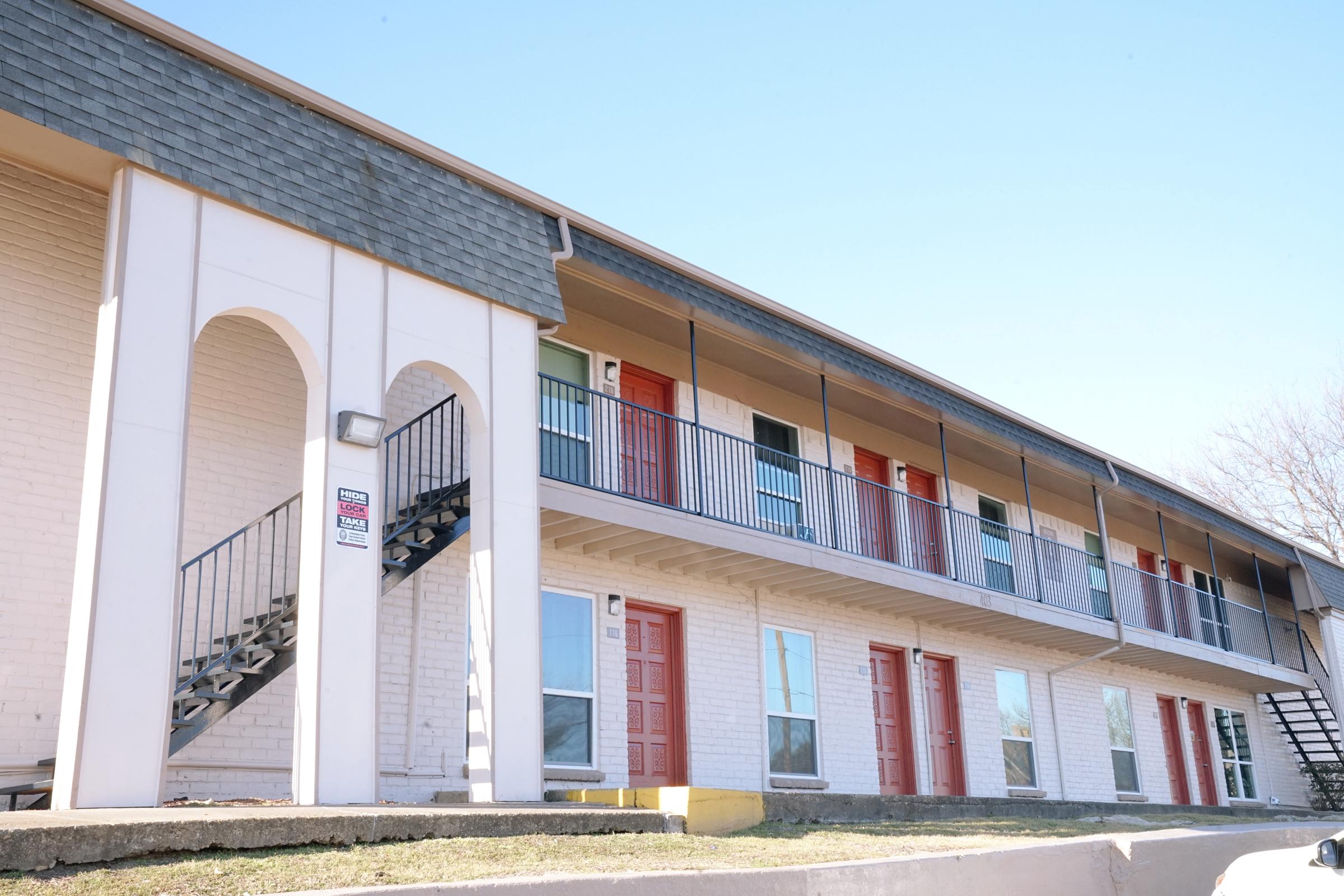 A two-story apartment building with a stairway on the left side. The building features multiple balconies with doors in various colors, including red and green. The exterior is light-colored brick with a gray roof, and the scene is set against a clear blue sky.