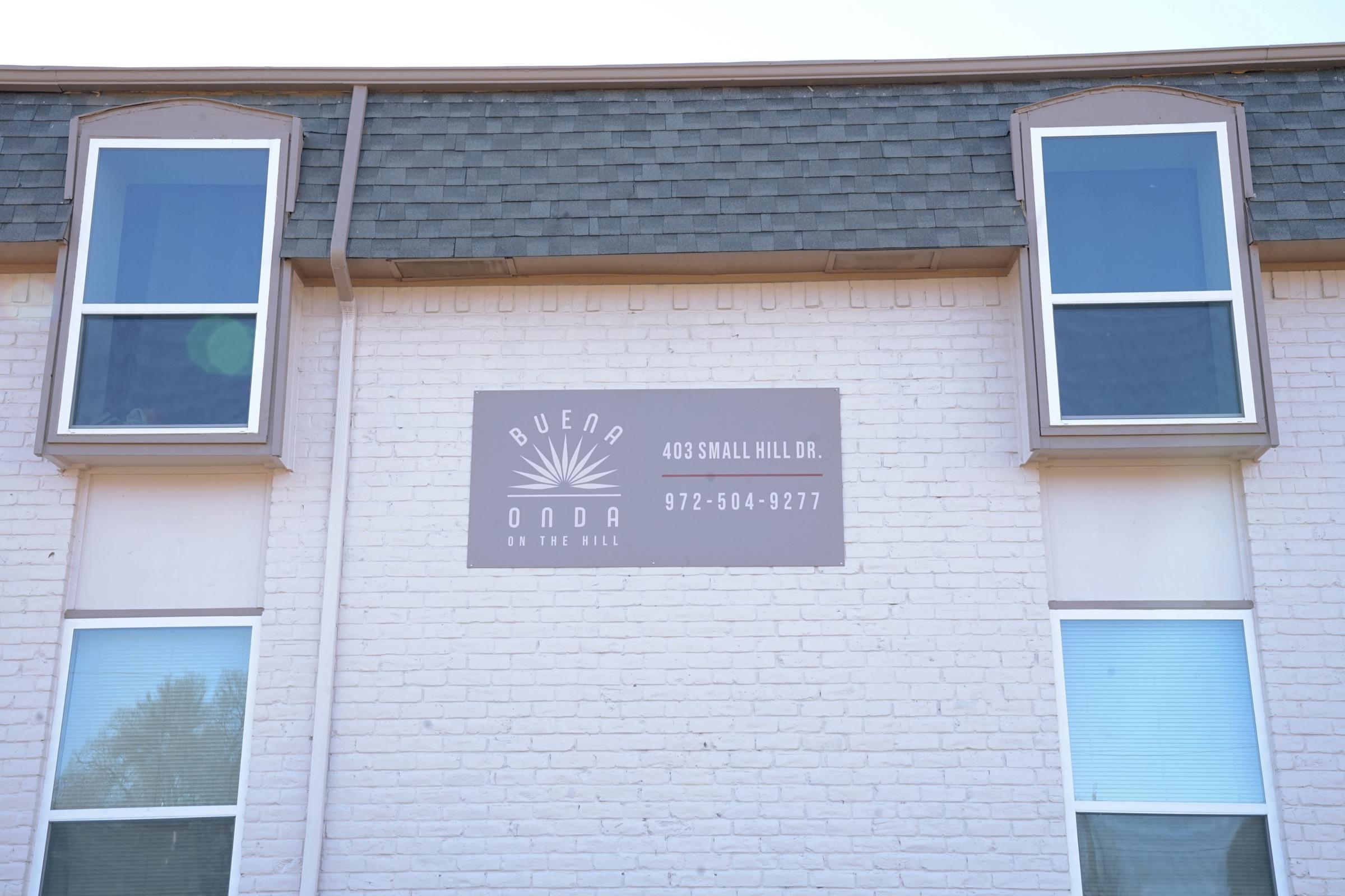 Sign on the side of a building with the text "BUENA ONDA" and the address "403 SMALL HILL DR. 972-504-9277." The building has two windows on the upper level and is painted in light colors, with a textured white brick wall.