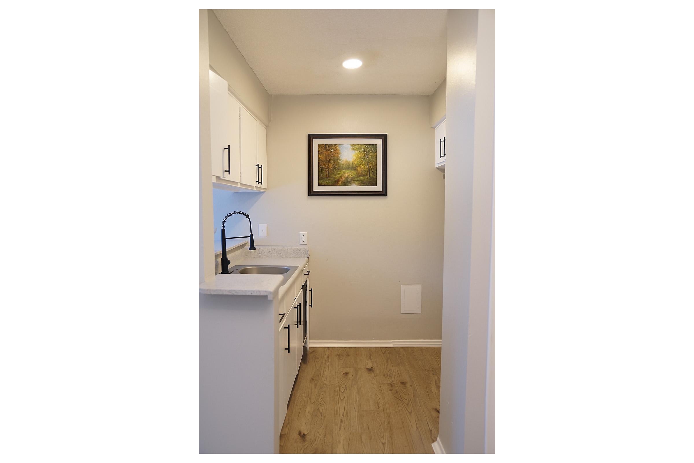 A compact kitchen with white cabinetry and a modern sink. There's a framed painting on the wall, and natural light from a ceiling fixture illuminates the space. The wooden floor adds warmth to the minimalist design. The room features a neutral color palette, creating a clean and inviting atmosphere.