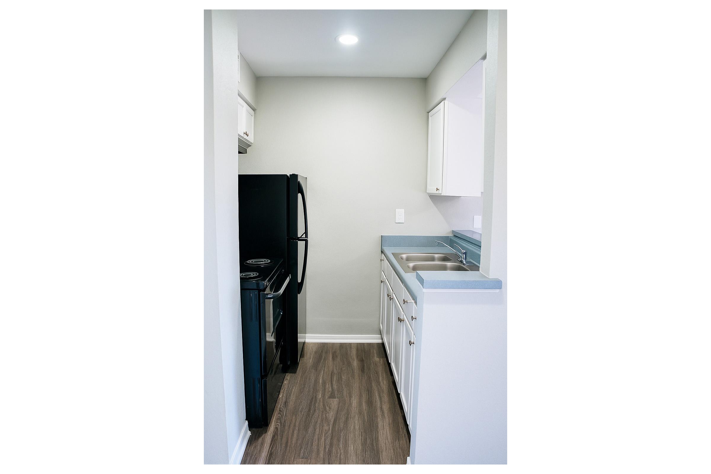 A modern kitchen featuring black appliances including a refrigerator, white cabinets, a double sink, and grey countertops. The space has light-colored walls and a hardwood floor, providing a clean and contemporary look. Bright overhead lighting enhances the area’s spacious feel.