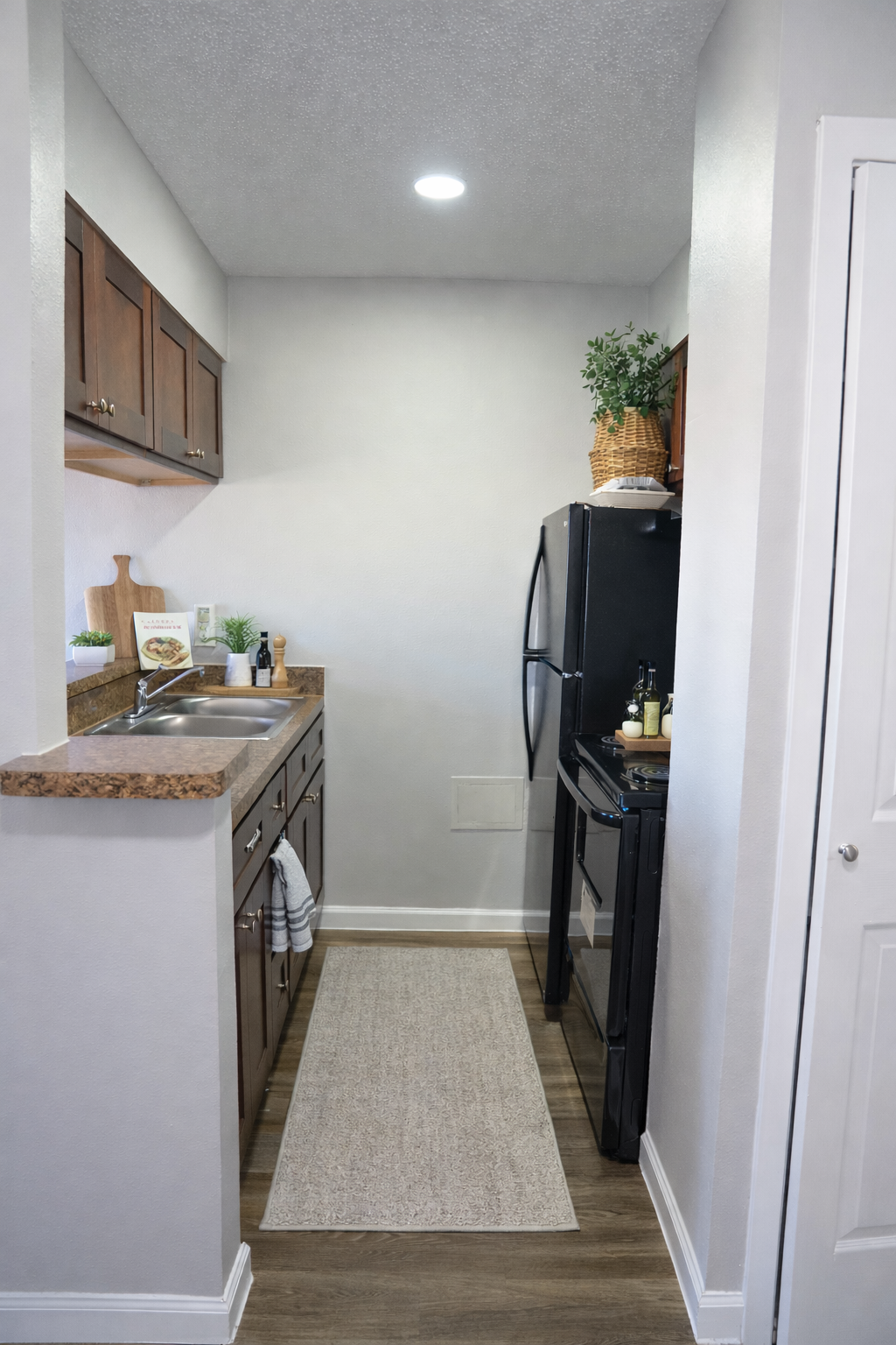 A small, modern kitchen featuring dark wood cabinetry, a stainless steel refrigerator, and an oven. The countertop is adorned with a cutting board and small plants. A neutral-colored runner rug extends along the floor, leading to a white door on the right, with soft lighting illuminating the space.