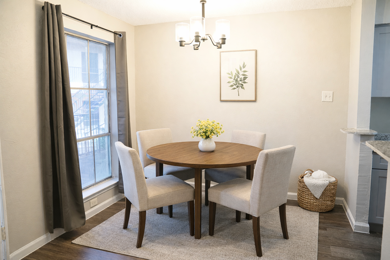 A cozy dining area featuring a round wooden table surrounded by four light-colored upholstered chairs. A vase of yellow flowers sits in the center of the table. The room has a warm, neutral color palette, with a large window letting in natural light and a decorative wall hanging. A woven basket is visible nearby.