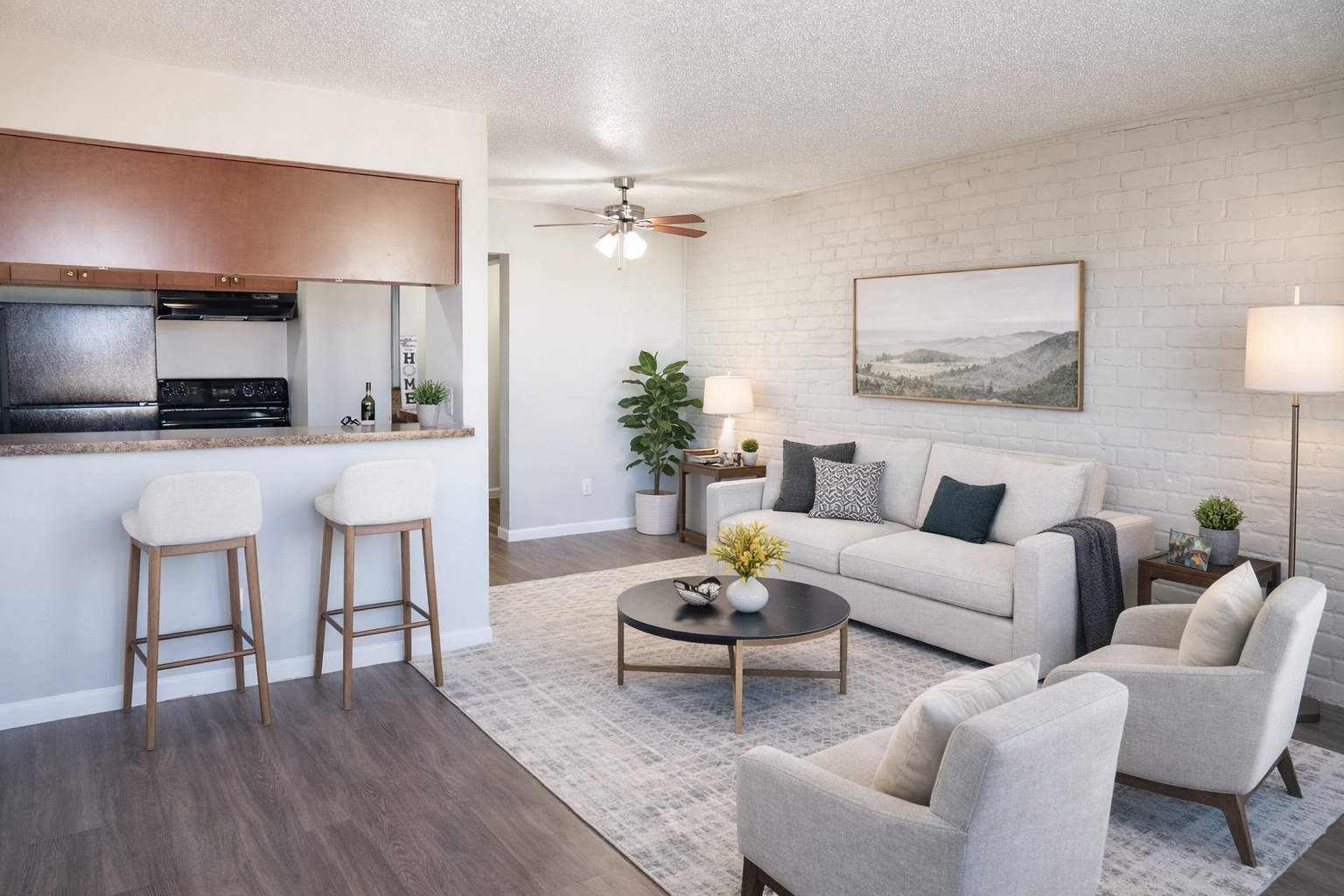 A bright and modern living space featuring a light-colored sofa, two armchairs, and a coffee table on a beige rug. In the background, there's a kitchen bar with two stools, and potted plants add a touch of greenery. A large landscape painting hangs on the wall, complementing the cozy atmosphere.