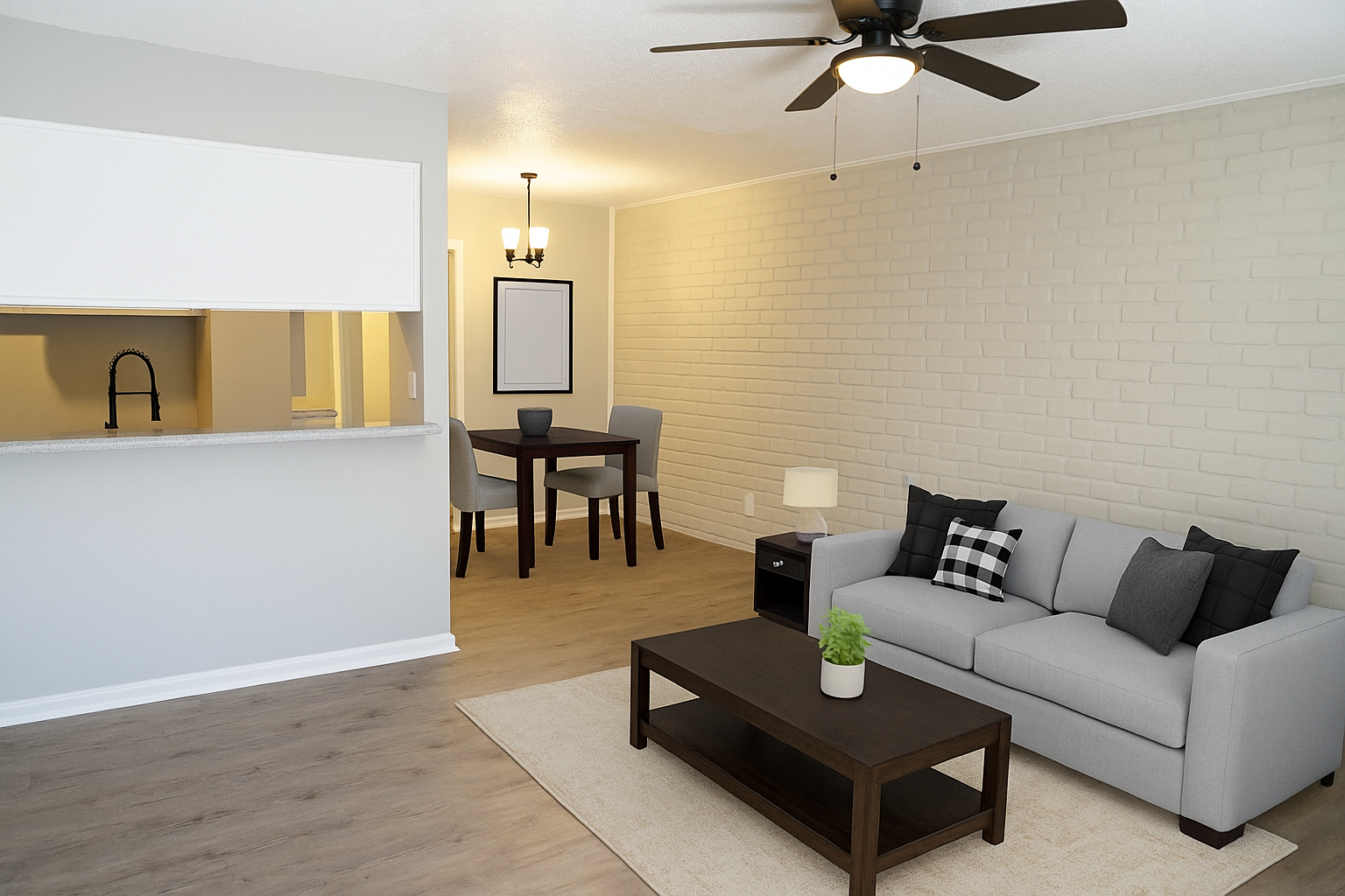 A modern, minimalist living room featuring a gray sofa with decorative pillows, a wooden coffee table, and a light-colored rug. In the background, there's a dining area with a small table and two chairs, and a kitchenette with a bar counter. The walls are painted in soft colors, and there's a ceiling fan.