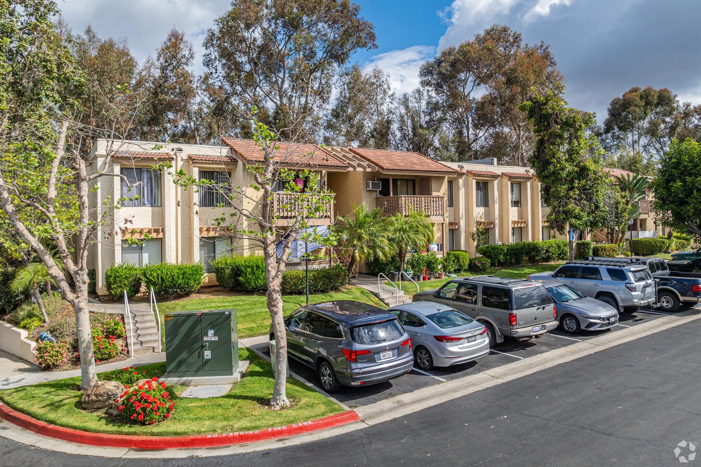 A residential apartment complex with multiple units surrounded by greenery. Parking spaces filled with vehicles line the front. There are trees and shrubs in the landscaping, and the sky is partly cloudy, suggesting a sunny day. The building features balconies and a well-maintained exterior.