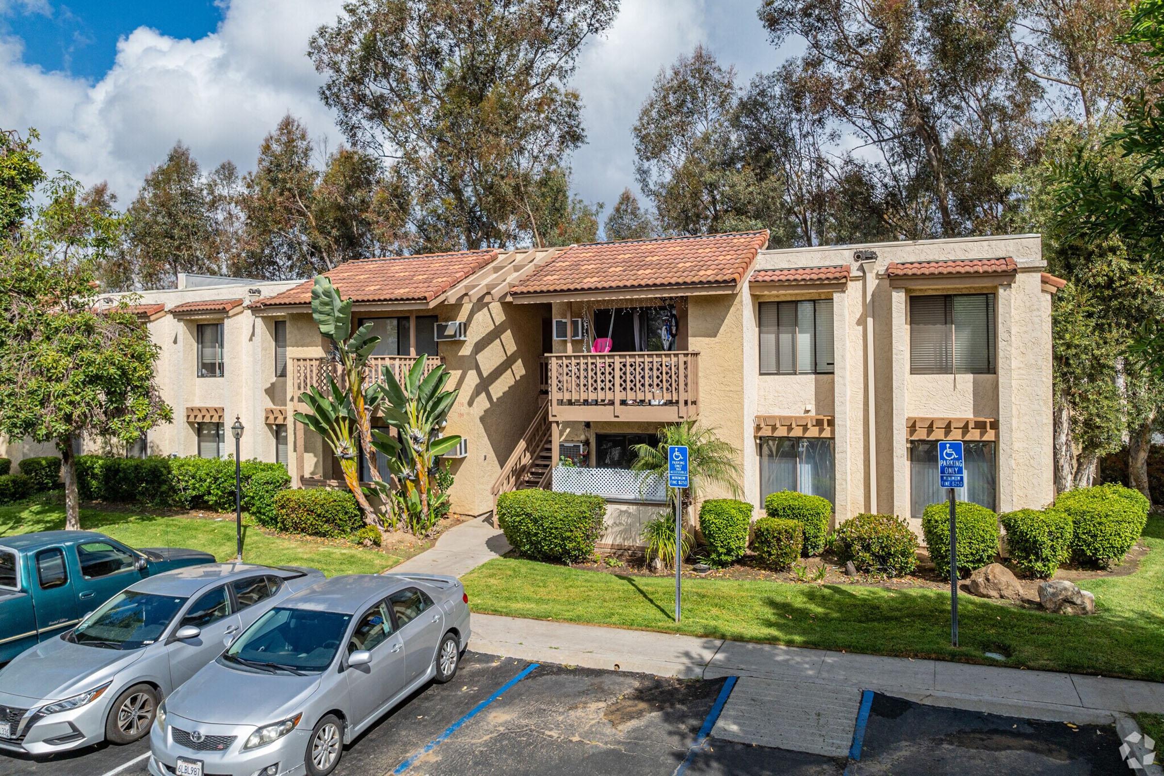 A view of a two-story apartment complex surrounded by trees and greenery. The building features balconies and is set against a partly cloudy sky. In the foreground, several parked cars are visible along a paved parking area, with accessible parking spaces indicated.