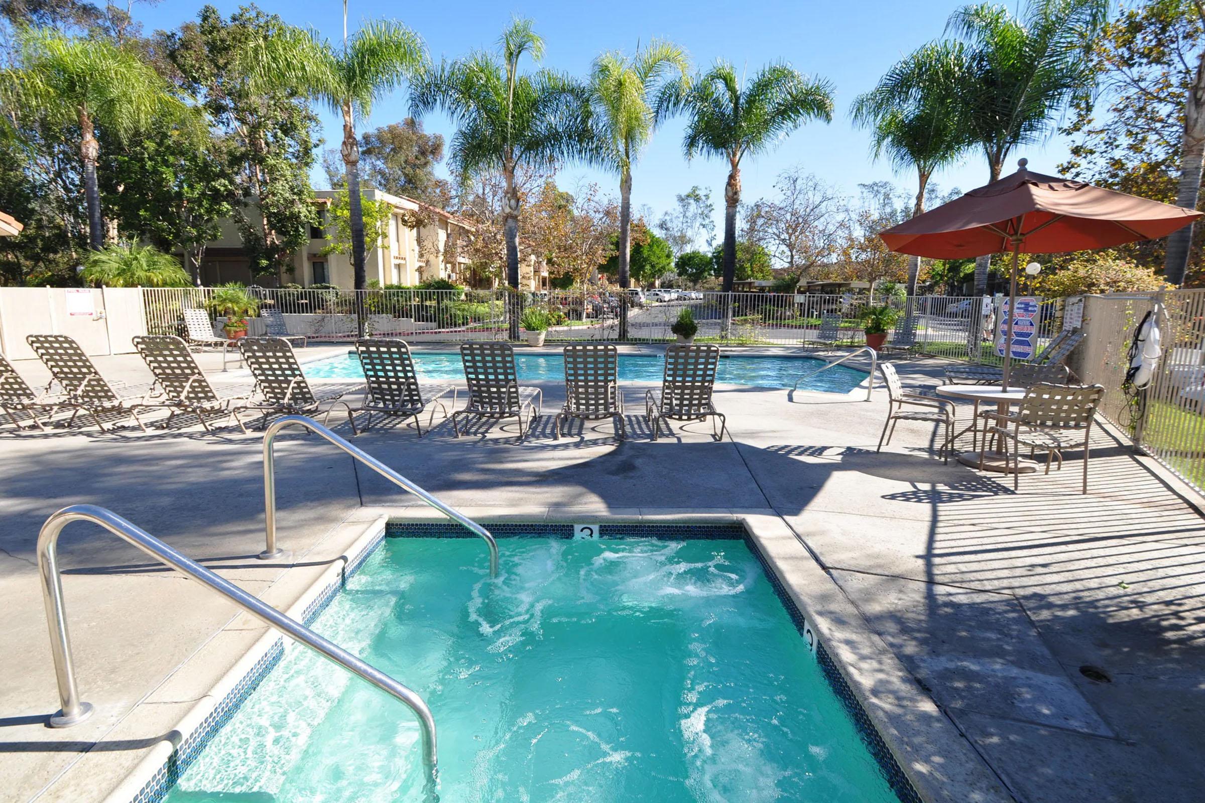 A brightly lit outdoor pool area featuring two swimming pools, one with spa jets. Surrounding the pools are several lounge chairs and a large umbrella providing shade. Tall palm trees and landscaped areas enhance the serene environment, making it an inviting space for relaxation.