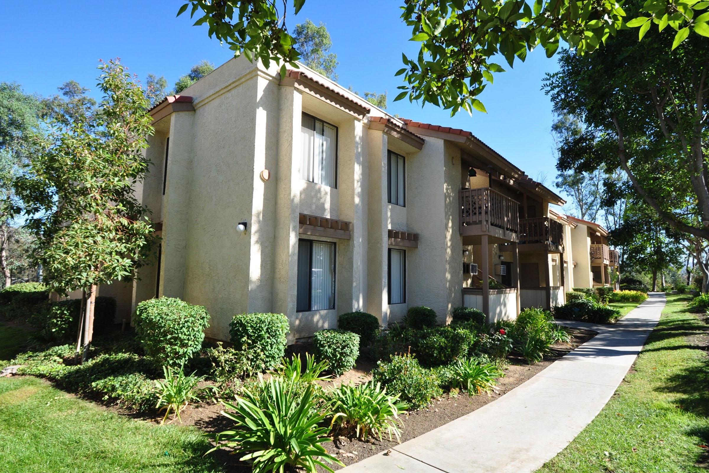 A well-maintained, two-story apartment building surrounded by lush greenery. The building features a light-colored exterior with balconies on the upper level. A pathway lined with shrubs and plants leads to the entrance, set against a clear blue sky. Sunlight casts a warm glow on the structure.