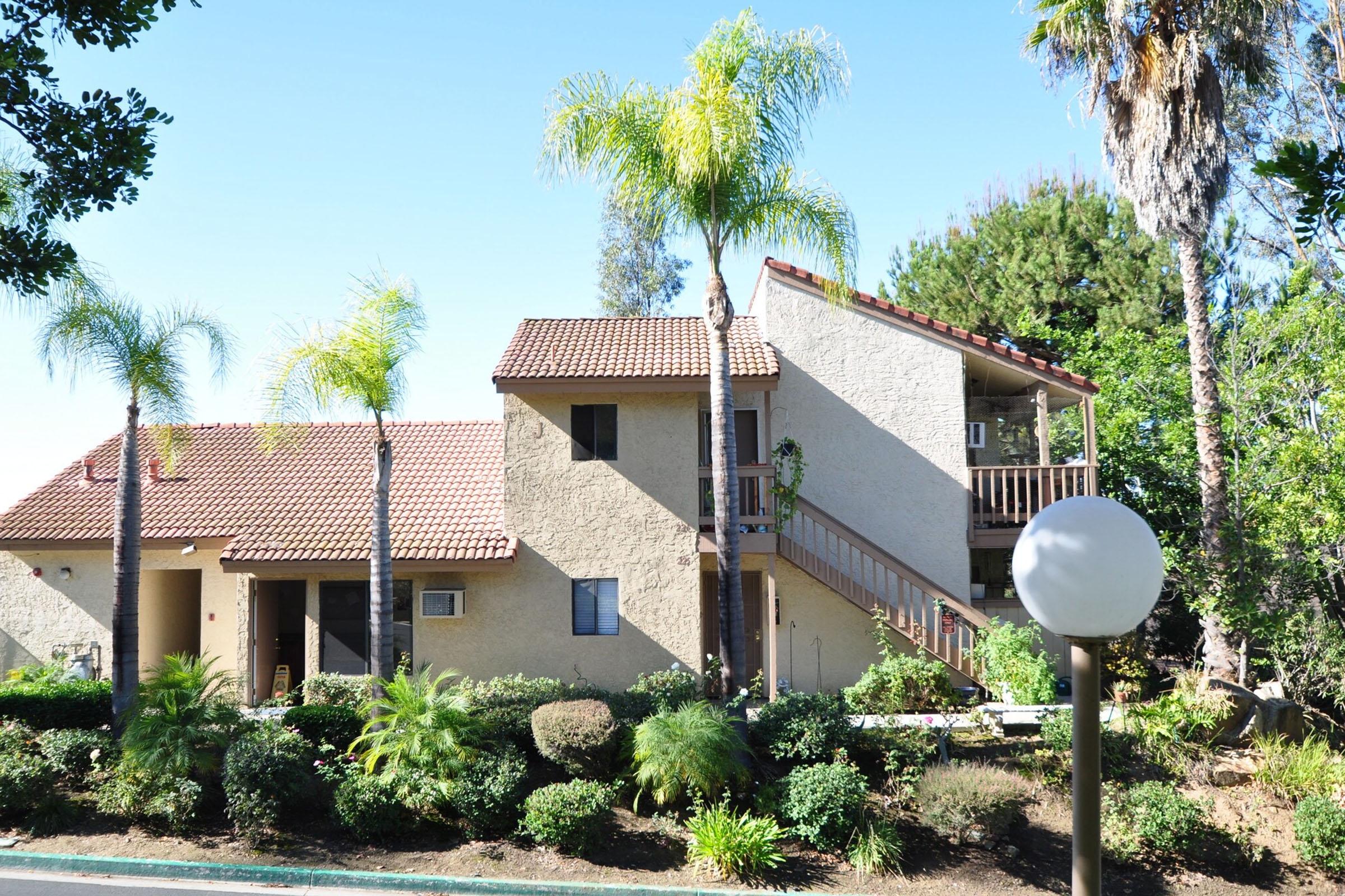 A modern two-story building with a textured beige exterior and a red tiled roof, surrounded by lush greenery and palm trees. There is a landscaped area in the foreground with various shrubs and plants, and a lamp post near the path leading to the entrance. The sky is clear and blue.