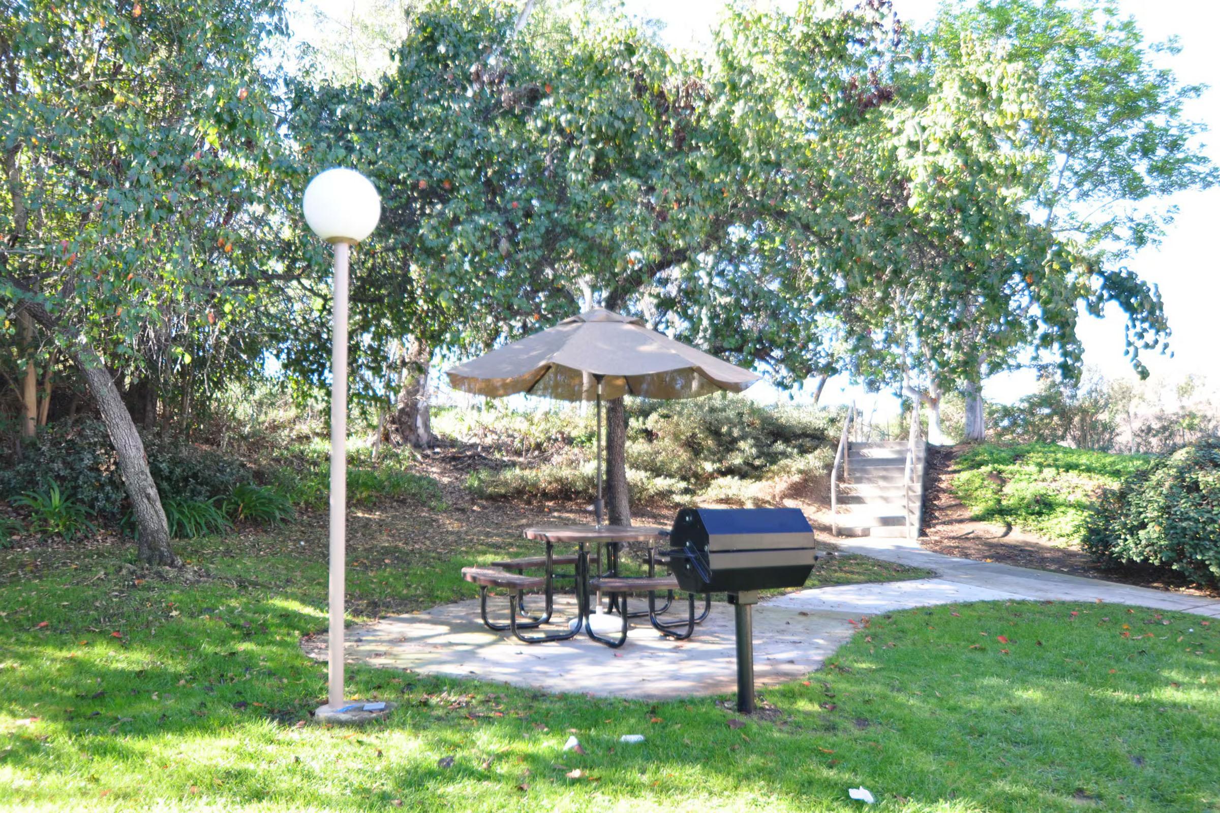 A tranquil picnic area in a park featuring a shaded table with benches beneath a large umbrella, a black grill nearby, and a pathway leading into the greenery. There is also a lamp post illuminating the space, surrounded by trees and well-maintained grass.