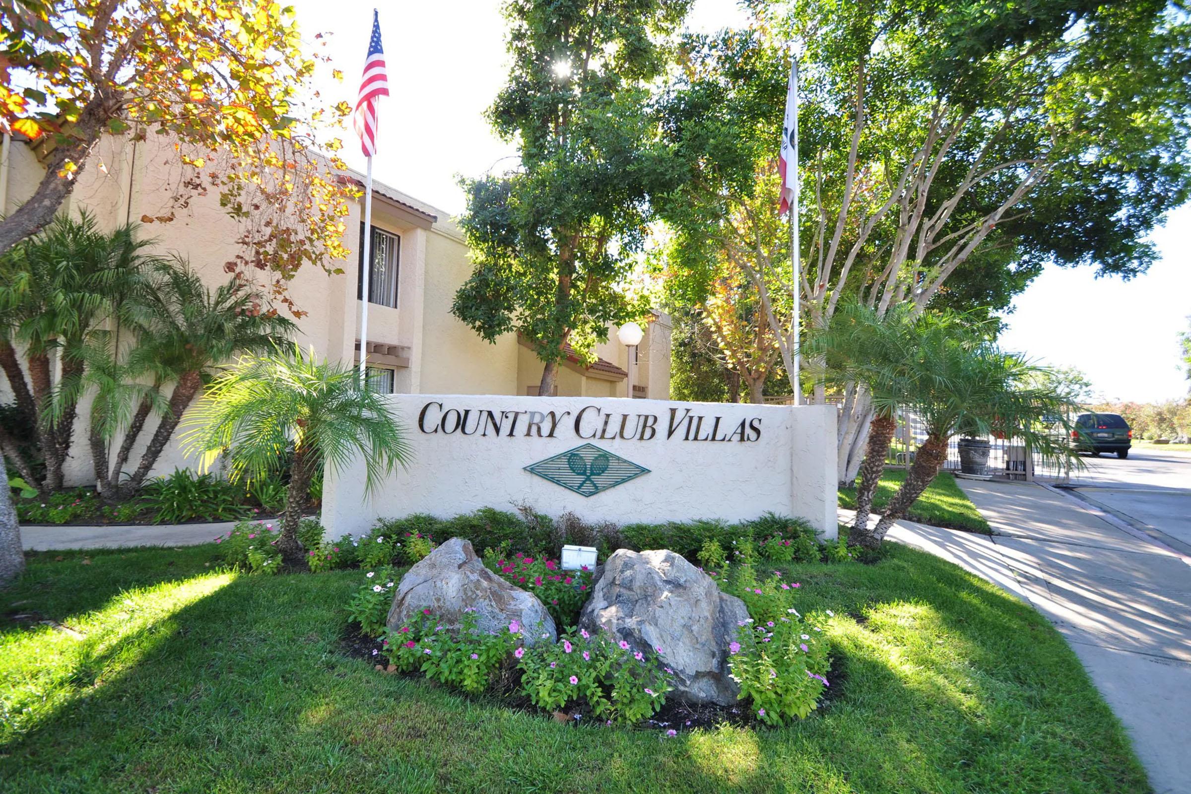 Sign for Country Club Villas featuring lush landscaping, including palm trees and colorful flowers, with the entrance of the building visible in the background. American and Texas flags are displayed. The scene conveys a welcoming atmosphere in a residential area.