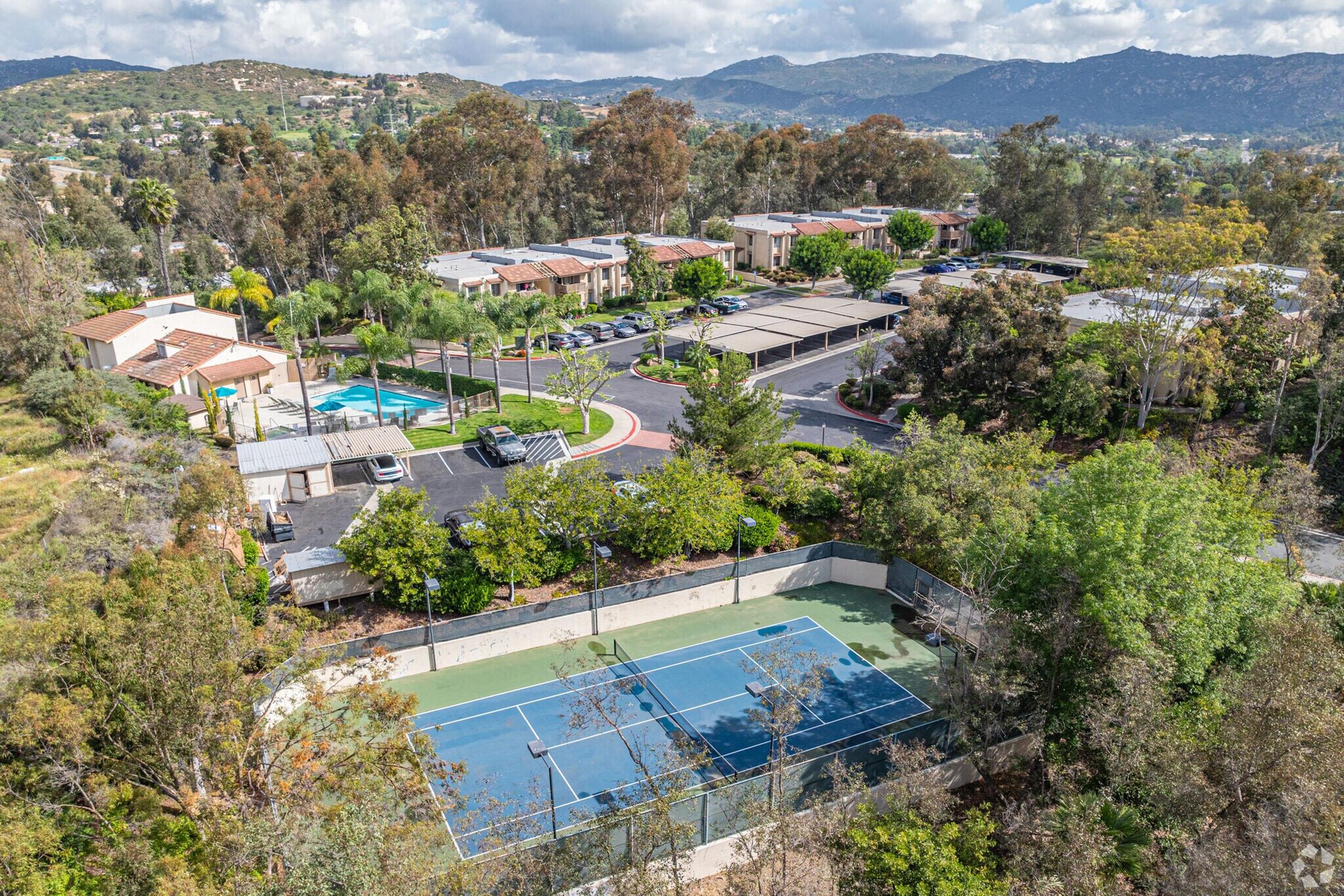 An aerial view of a residential area featuring a tennis court surrounded by trees. In the background, there are several homes with red-tiled roofs, a swimming pool, and landscaped gardens. The scene is set against rolling hills under a partly cloudy sky.