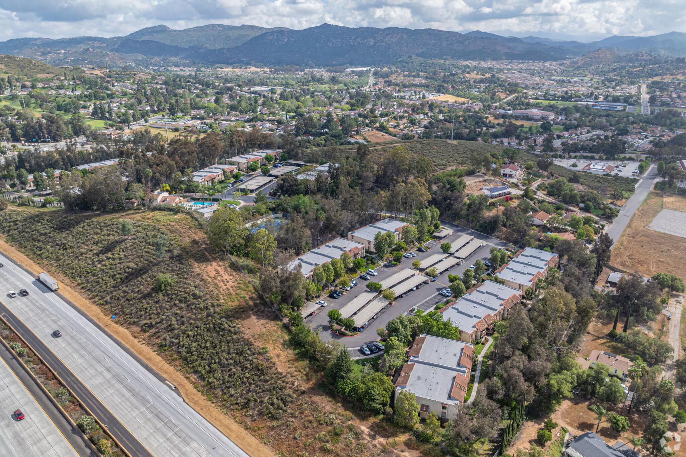 Aerial view of a suburban area featuring low-rise residential buildings and green spaces, bordered by a highway. The landscape includes rolling hills and a mix of urban development with natural scenery, showcasing a combination of homes, trees, and distant mountains under a partly cloudy sky.