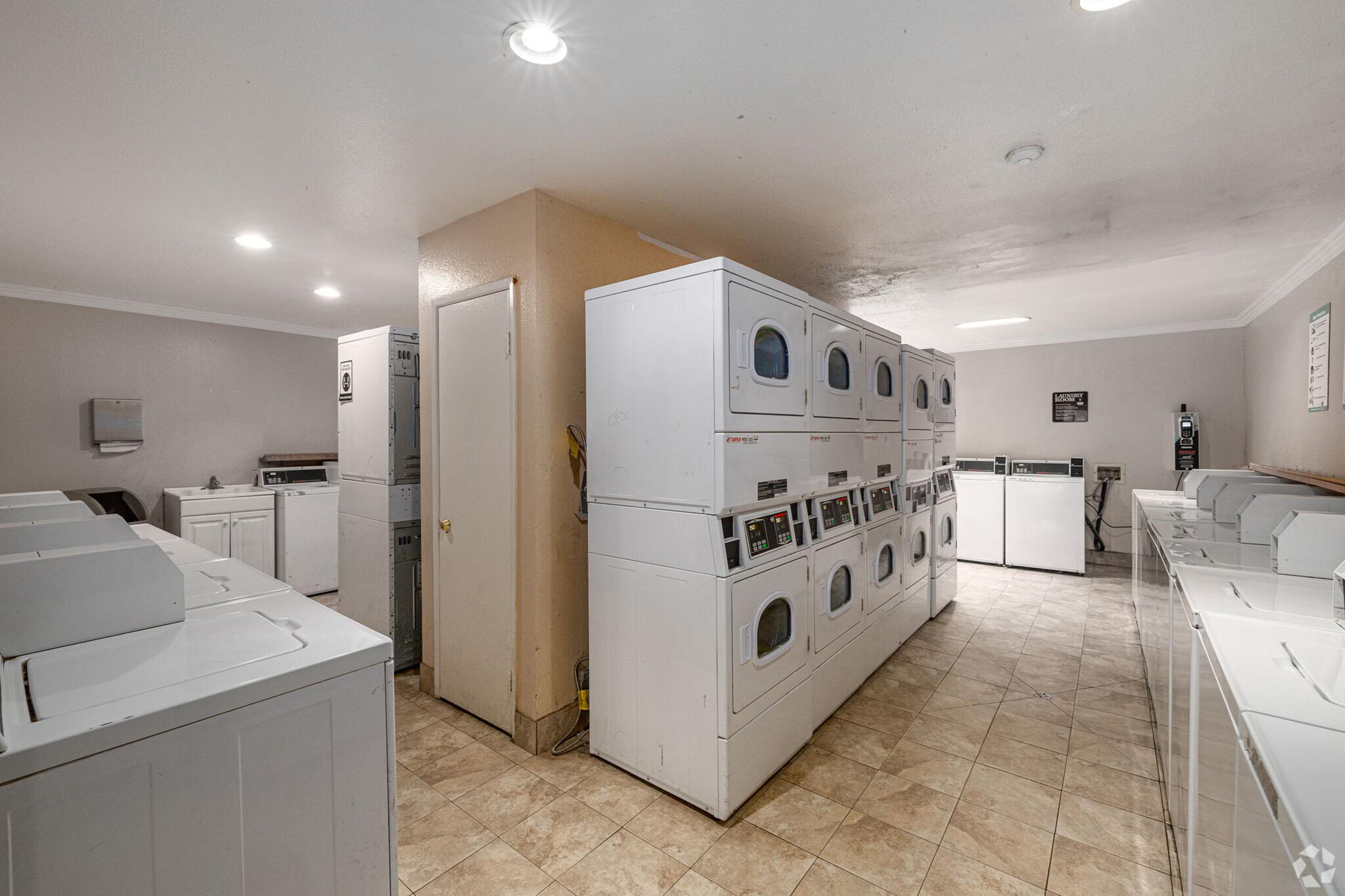 A clean laundry facility featuring rows of white washing machines and dryers. The room is well-lit with overhead lights, and there is a door visible on one side. The tiled floor adds to the neat appearance, and the walls are painted a light color. Various laundry supplies and a folding area are present.