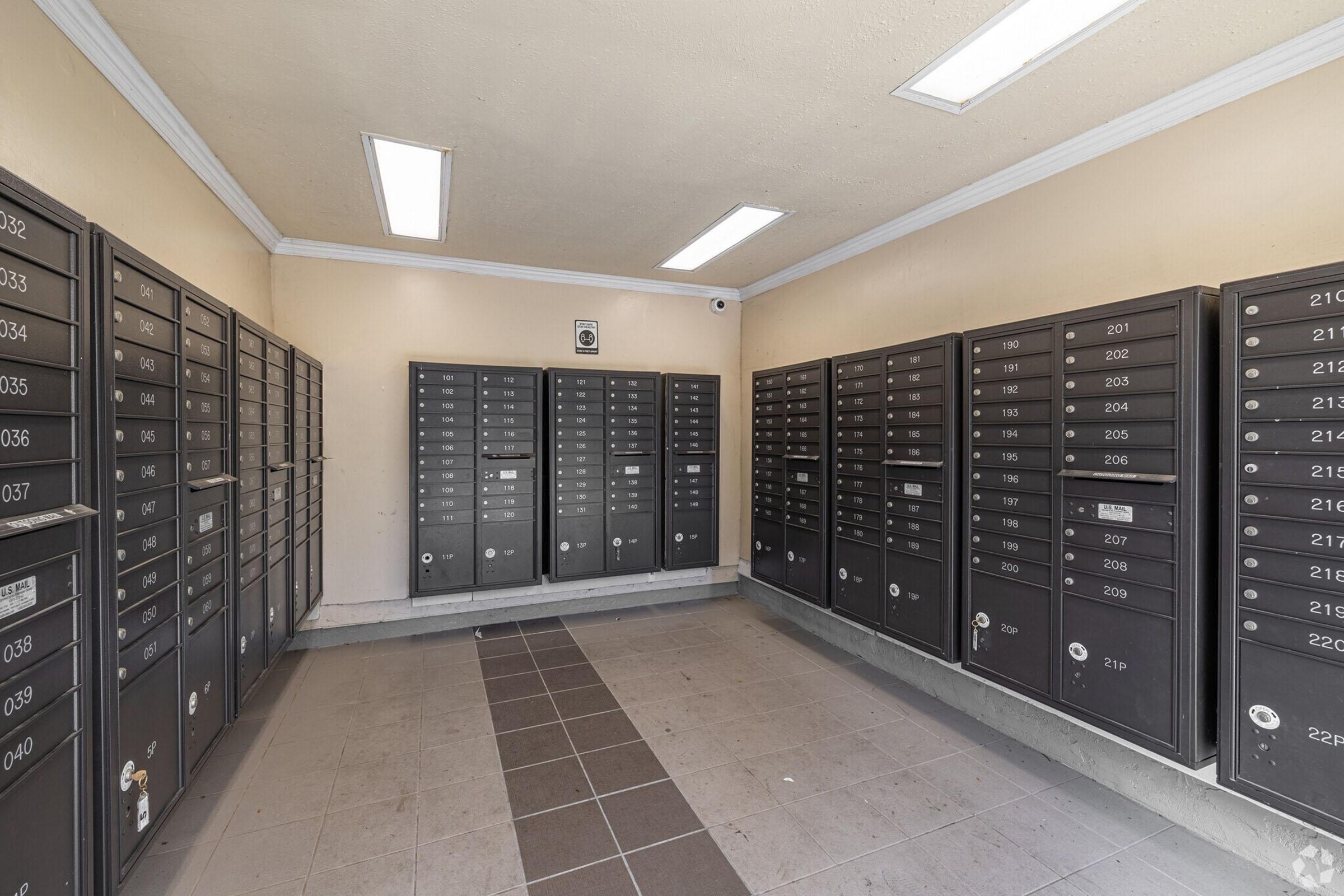 Interior view of a postal mailroom with rows of black mailboxes arranged against the walls. Bright overhead lights illuminate the space, which has a neutral-colored wall and tiled floor. The layout is orderly, with each mailbox numbered and accessible.