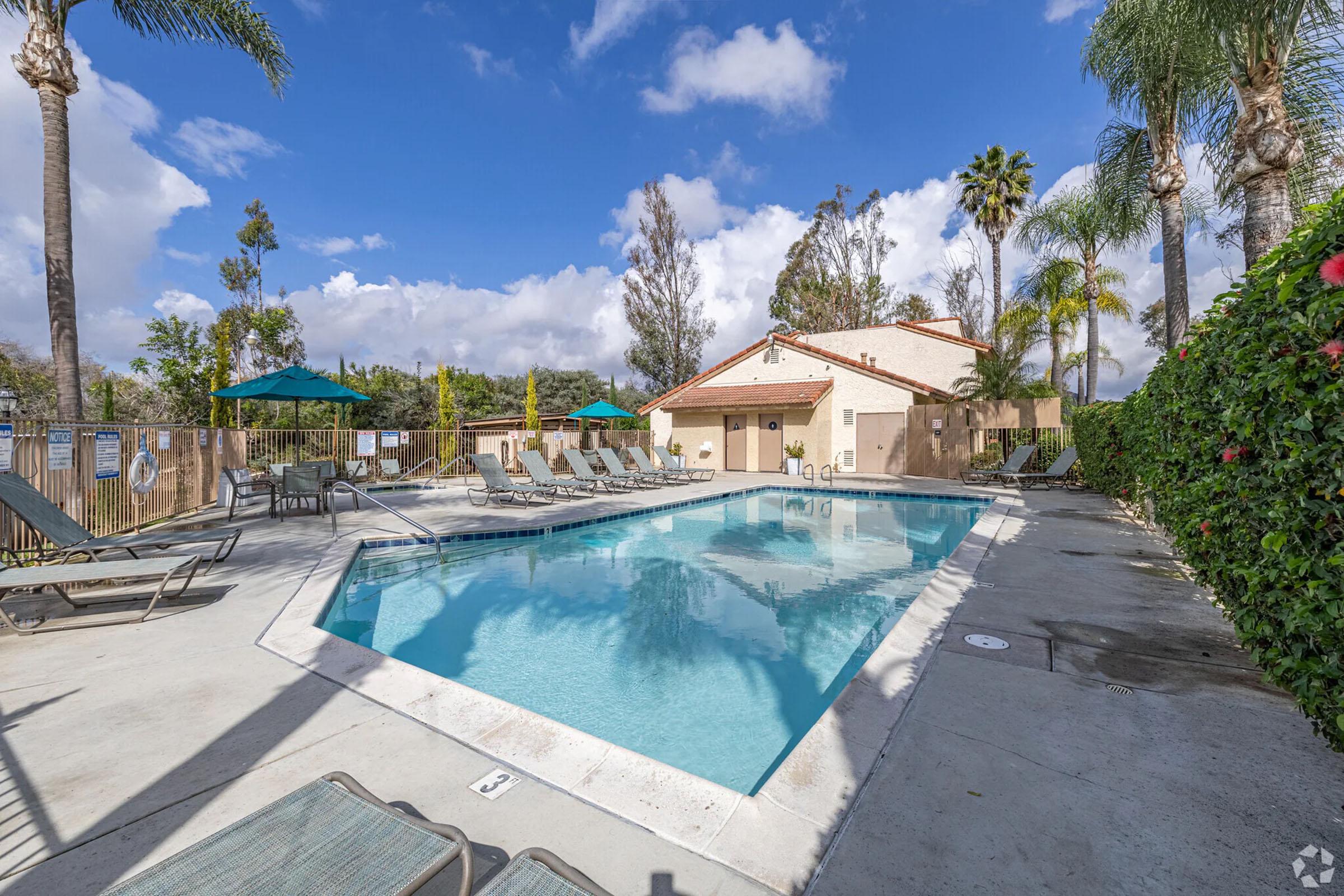 A clear, inviting swimming pool surrounded by sun loungers, with palm trees and vibrant greenery in the background. A light-colored building is visible nearby, and the sky features some fluffy clouds, creating a pleasant setting for relaxation.