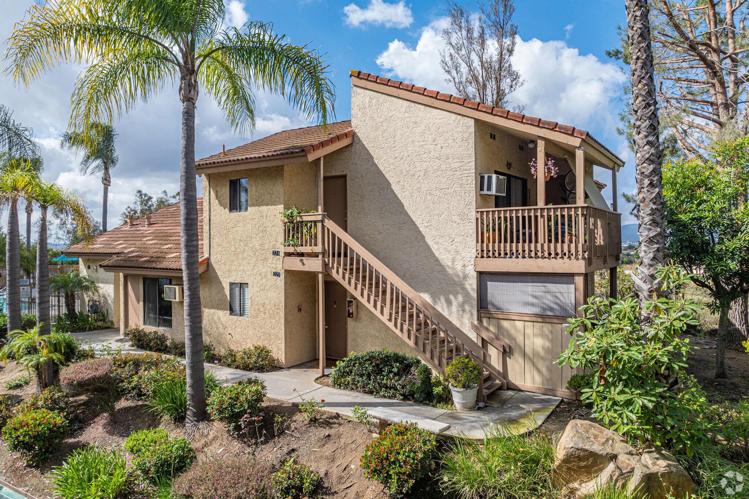 A two-story residential building with a beige exterior, red roof tiles, and a wooden staircase leading to the upper level. Lush greenery including palm and other plants is seen in the foreground. The sky is partly cloudy, creating a bright atmosphere.