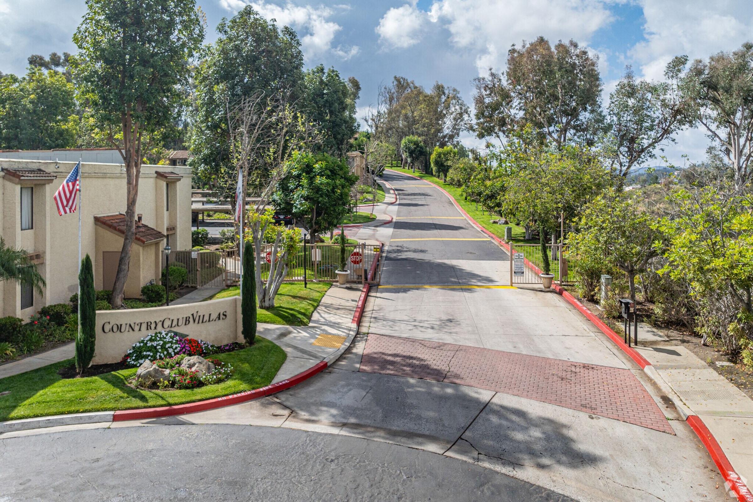 View of a residential area named "Country Club Villas," featuring a winding road lined with trees and landscaping. An American flag is visible on a building, and the scene is bright with blue skies and some clouds. The road curves gently upward, suggesting a suburban environment.