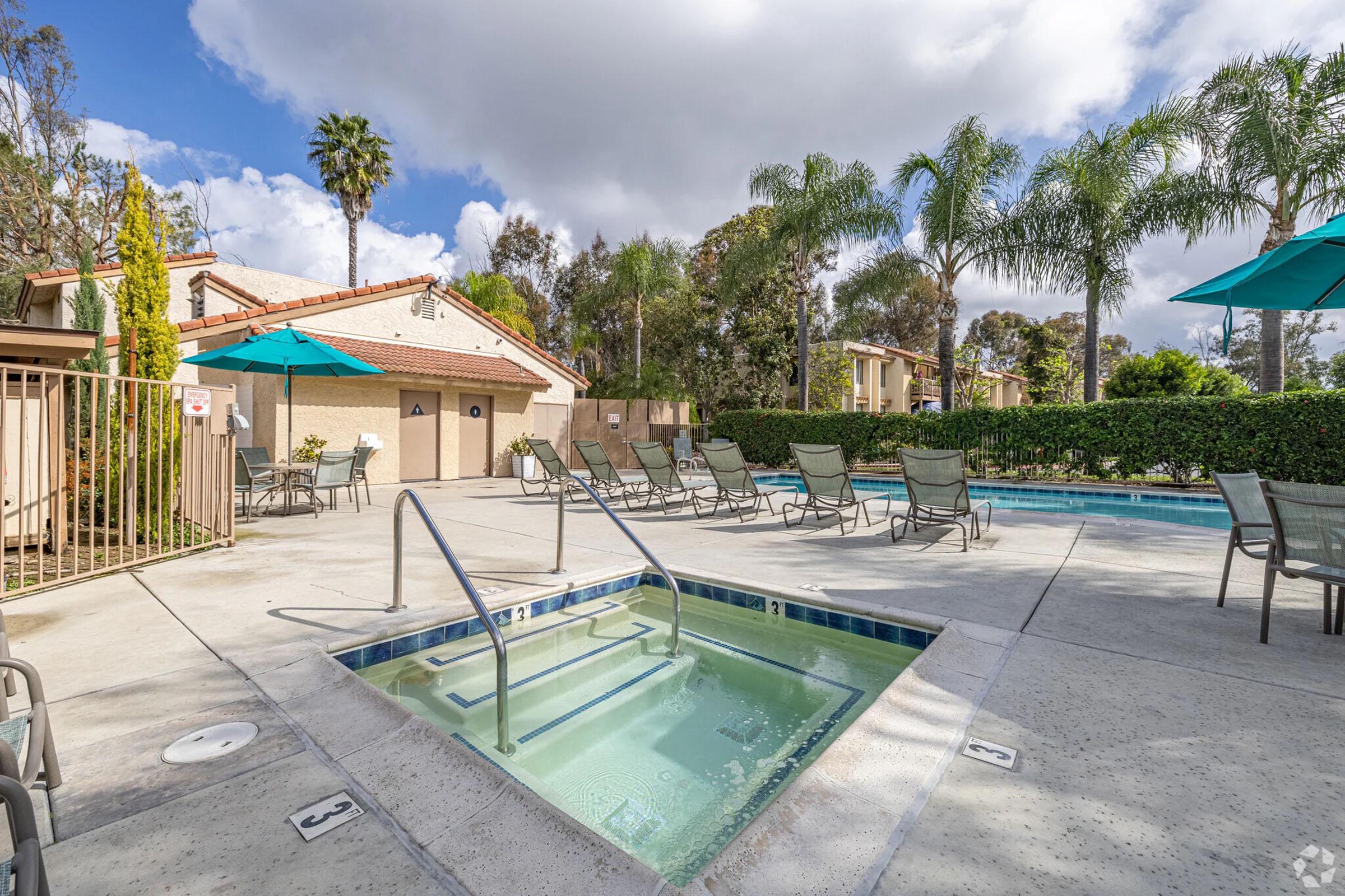 A view of a sunny pool area featuring a hot tub and swimming pool. Surrounding the pool are lounge chairs, palm trees, and umbrellas. In the background, there are residential buildings and landscaped greenery. The sky is partly cloudy, enhancing the outdoor atmosphere.