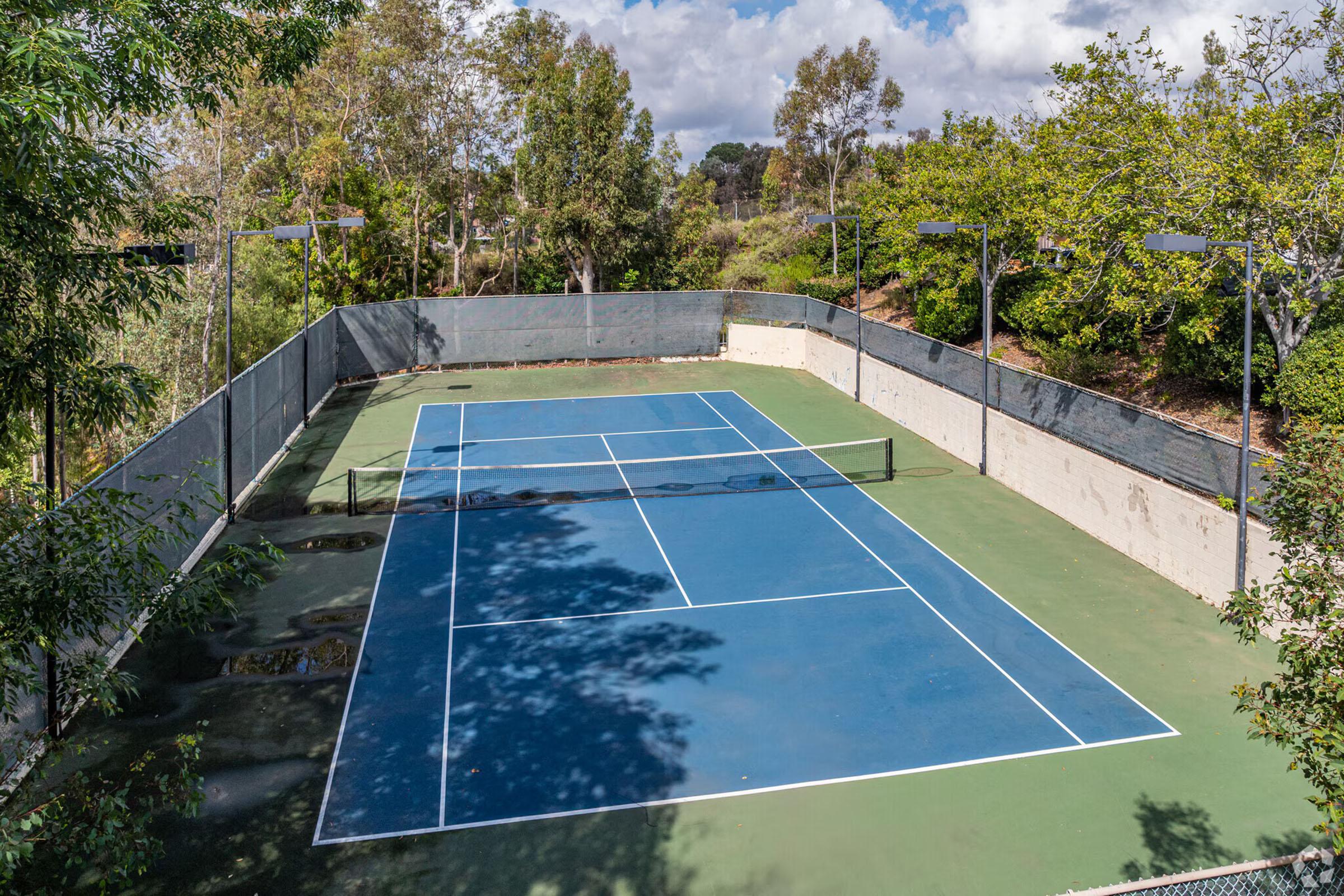 Aerial view of a tennis court surrounded by trees and greenery. The court features blue and green playing surfaces, with netting visible in the center. Sunlight casts shadows on the court, and there are floodlights installed along the perimeter. The atmosphere is calm and inviting for a game.