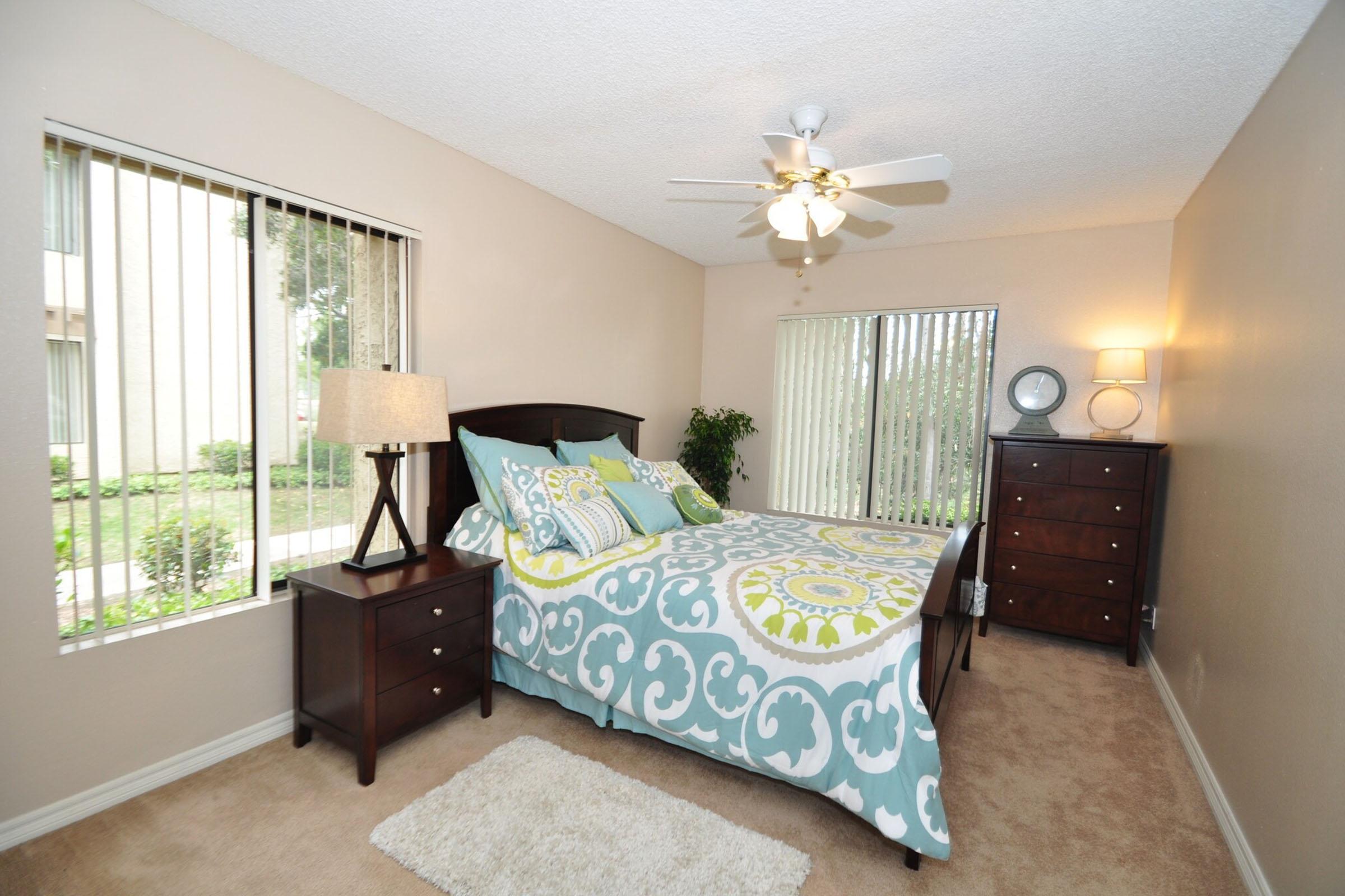 A cozy bedroom featuring a queen-sized bed with a colorful printed duvet, two nightstands with lamps, a dresser, and large windows with vertical blinds. The room has light beige walls and carpet, and a small area rug. A ceiling fan is present, and there is greenery visible outside the windows.