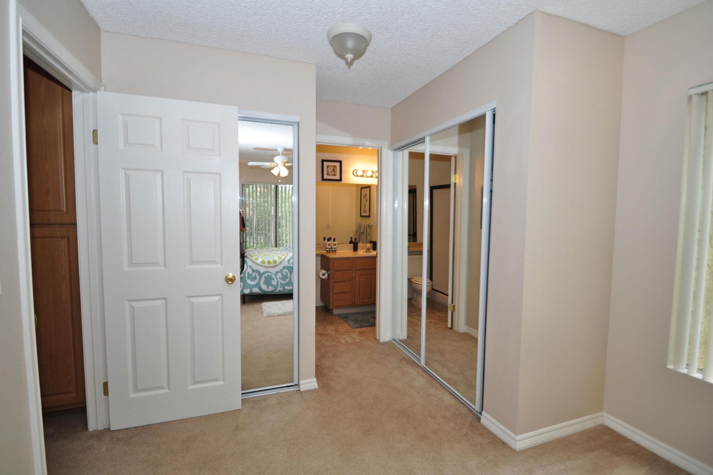A view of a light-colored room with a beige carpet, featuring a doorway leading to a bathroom and a bedroom. There are mirrored sliding closet doors on one wall, and a ceiling light fixture. Natural light comes in through windows with blinds, and the adjacent bathroom has a vanity and sink visible in the reflection.