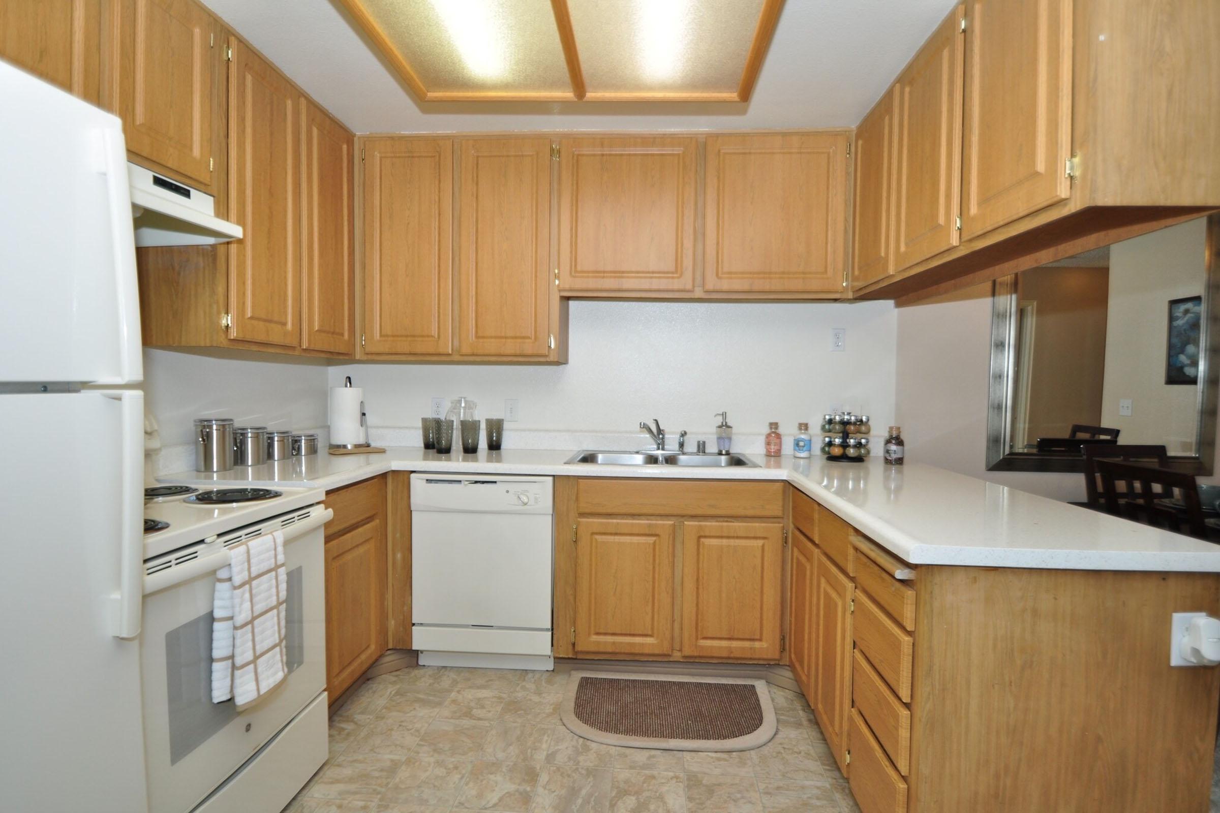 A modern kitchen featuring wooden cabinets, a white countertop, a double sink, a dishwasher, and an oven with a stovetop. The kitchen includes a refrigerator, a kitchen mat on the floor, and a shelf with various jars. The lighting is warm, enhancing the inviting atmosphere.