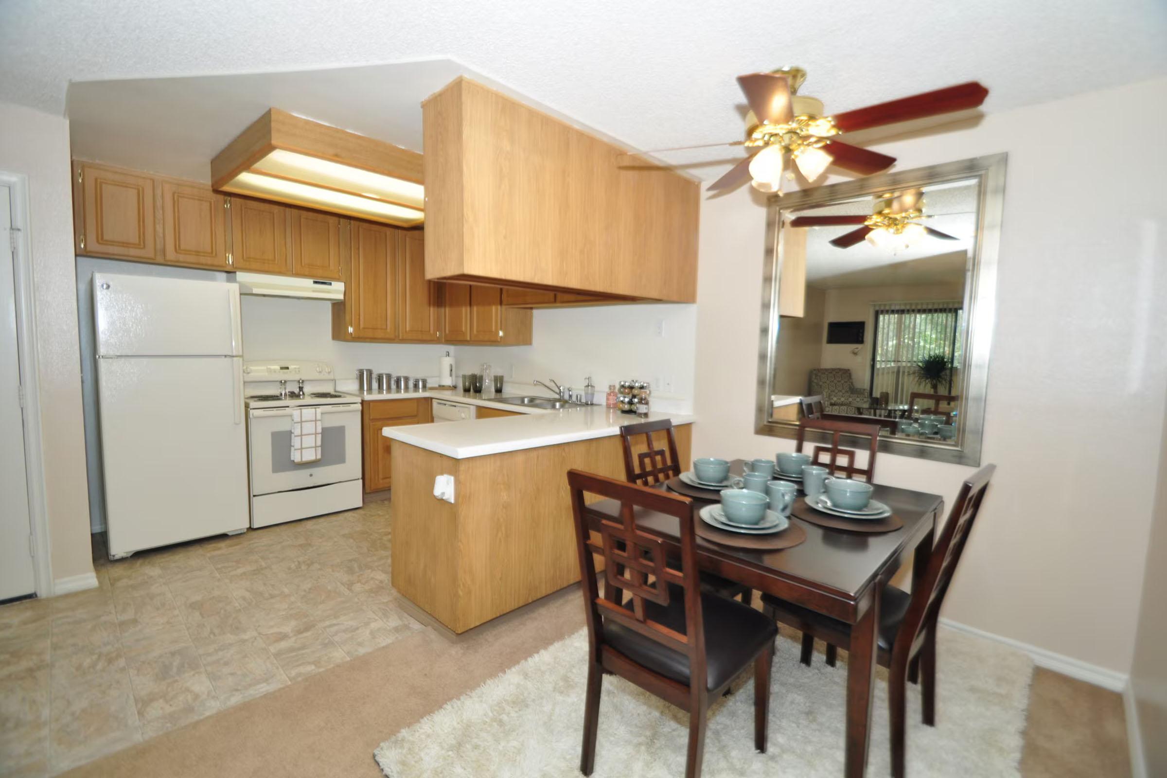 A modern kitchen with light wood cabinetry and a white countertop. The kitchen includes a gas stove and refrigerator. Adjacent is a dining area featuring a dark wood table set for four with teal dishware, and a ceiling fan with wooden blades. A large mirror reflects the space, enhancing openness.